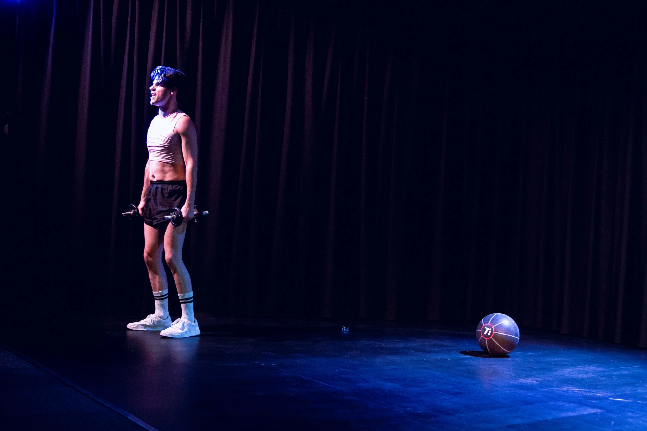 A person with short blue hair and a crop top holding dumbbells, standing on a stage in front of dark curtains, with a basketball on the floor to the right.