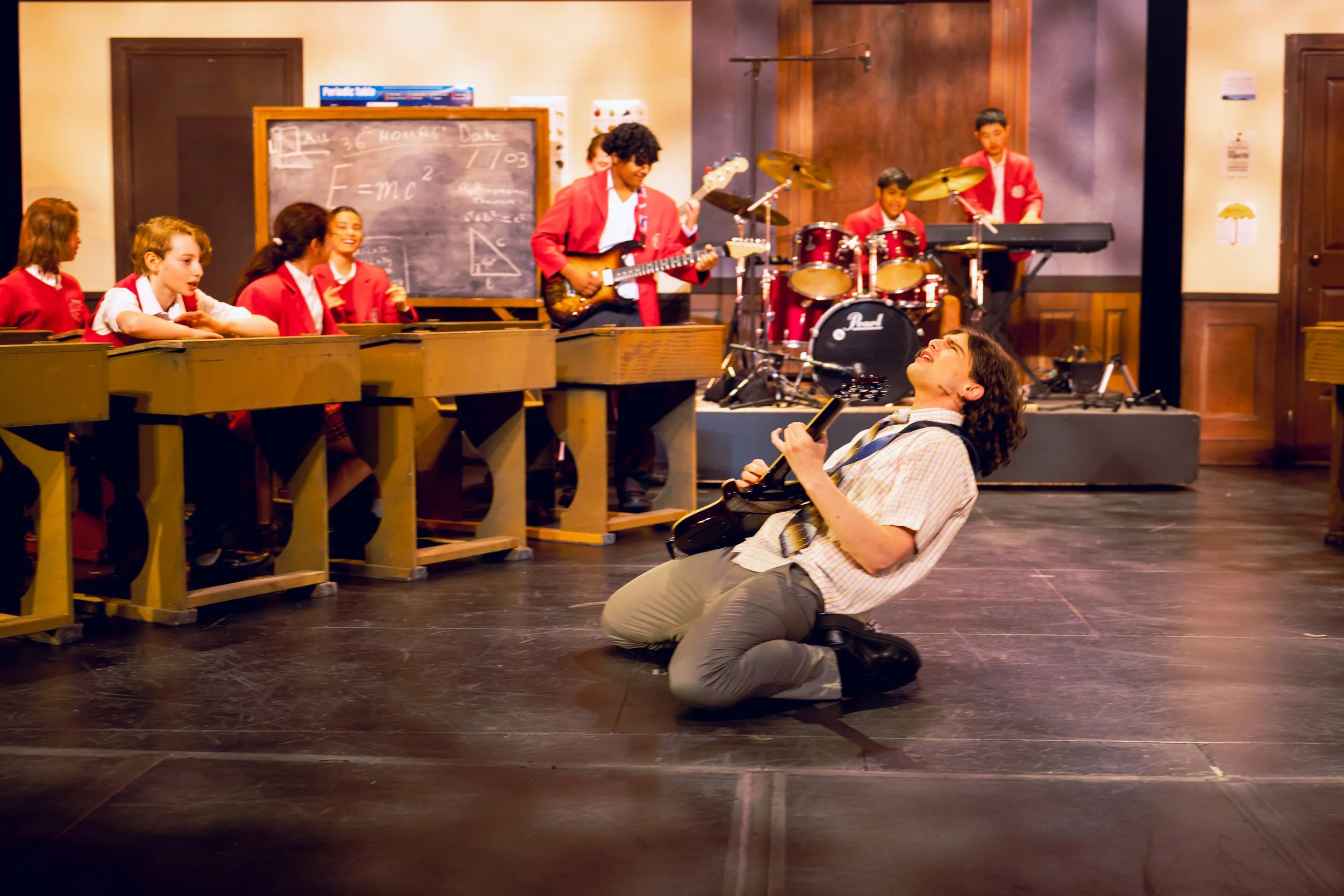 A music teacher kneeling on the floor playing air guitar while students in school uniforms watch a band perform on a stage in a classroom setting.