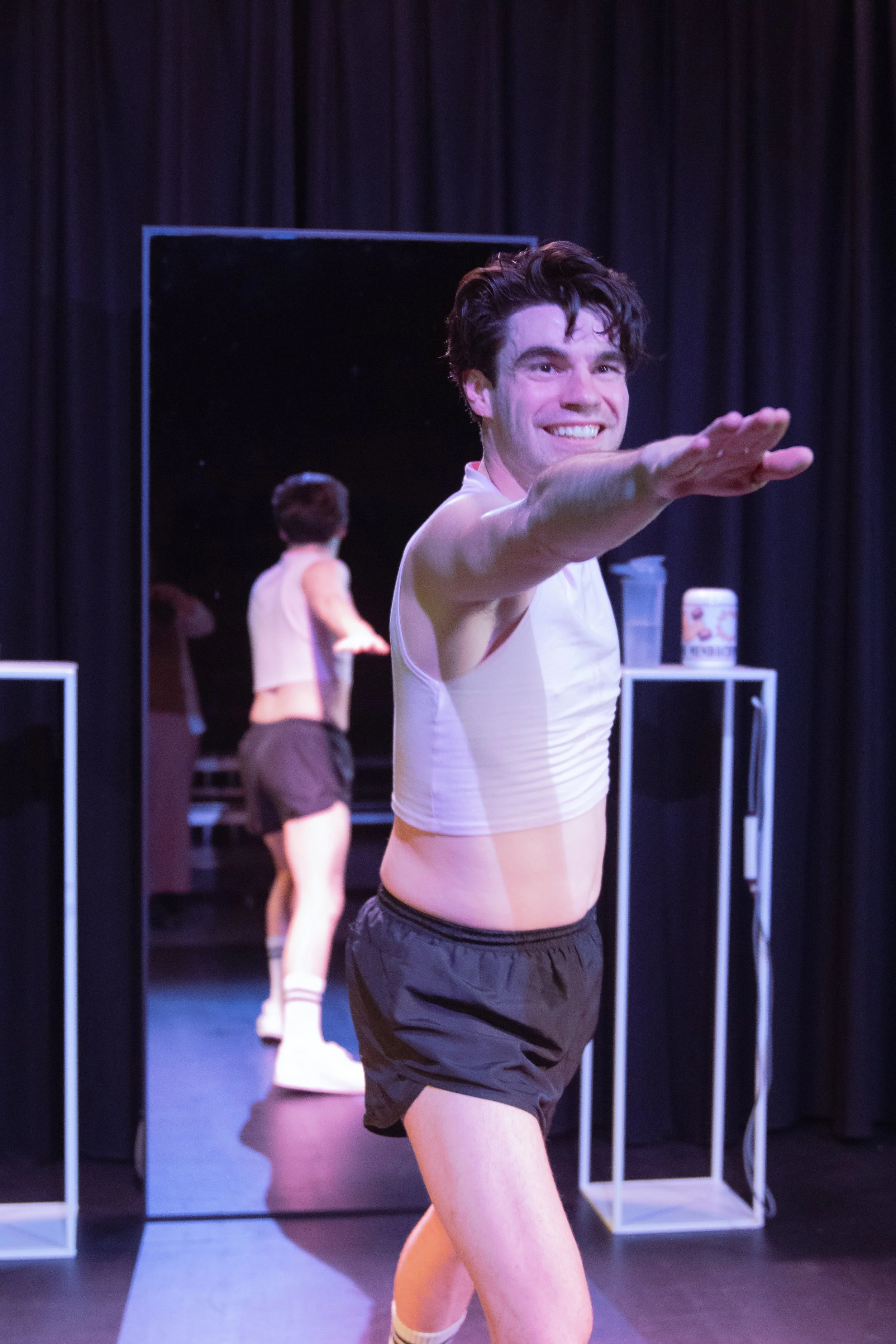 Man smiling in a white tank top and black shorts, reaching towards the camera, with a mirror and a woman in the background on stage.