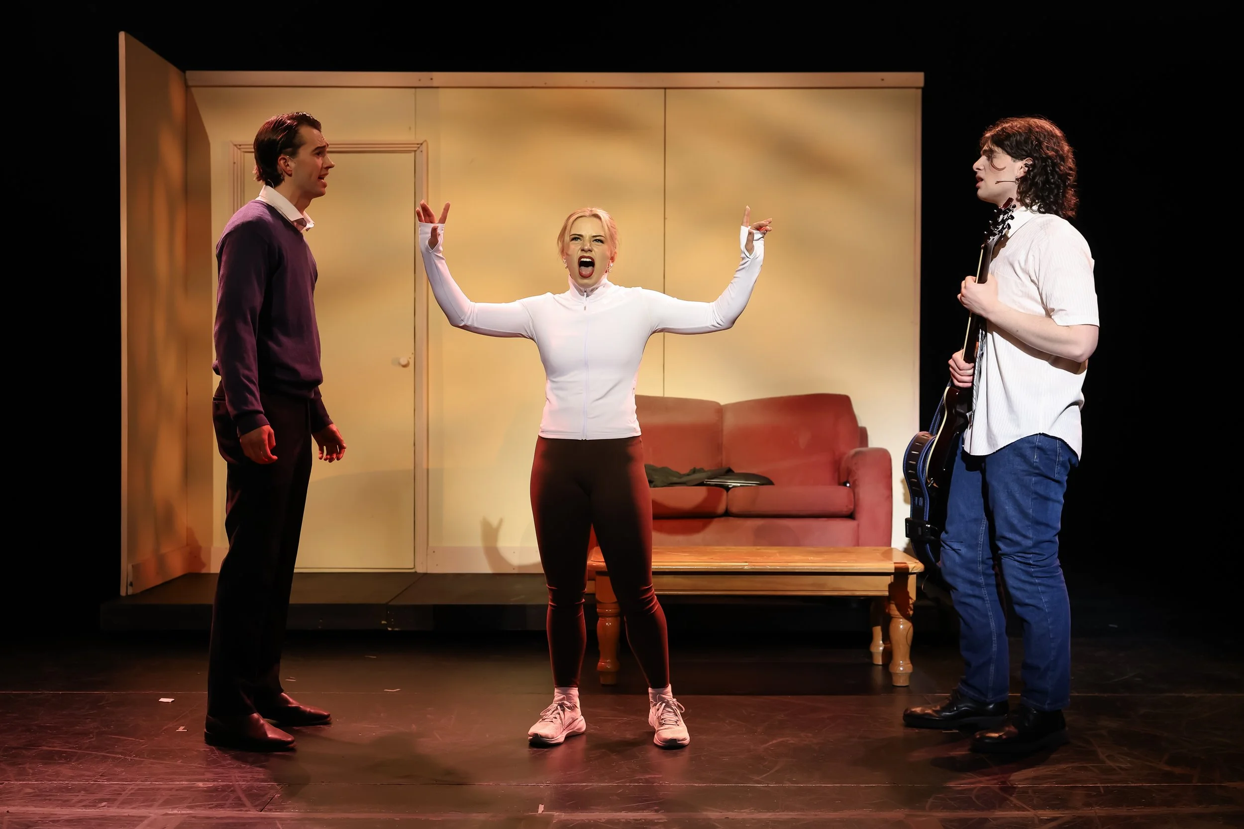 Three actors performing on a stage with a beige wall, a door, a sofa, and a coffee table in the background. The woman in the middle is shouting with arms raised, wearing a white zip-up top and black pants. The man on the left is facing her, wearing a