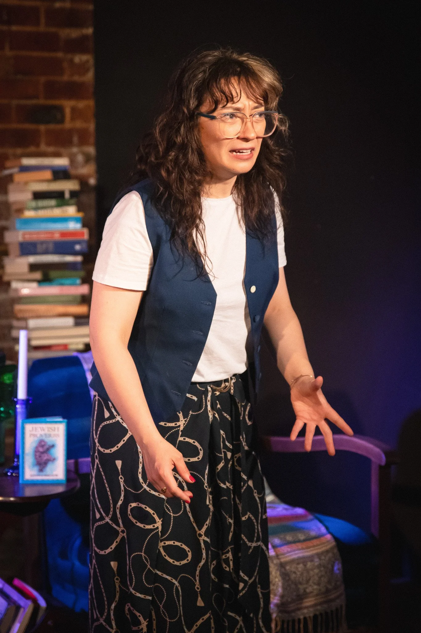 A woman with curly brown hair and glasses, wearing a white t-shirt, navy vest, and patterned black pants, appears to be performing on stage with a book and bookshelves in the background.