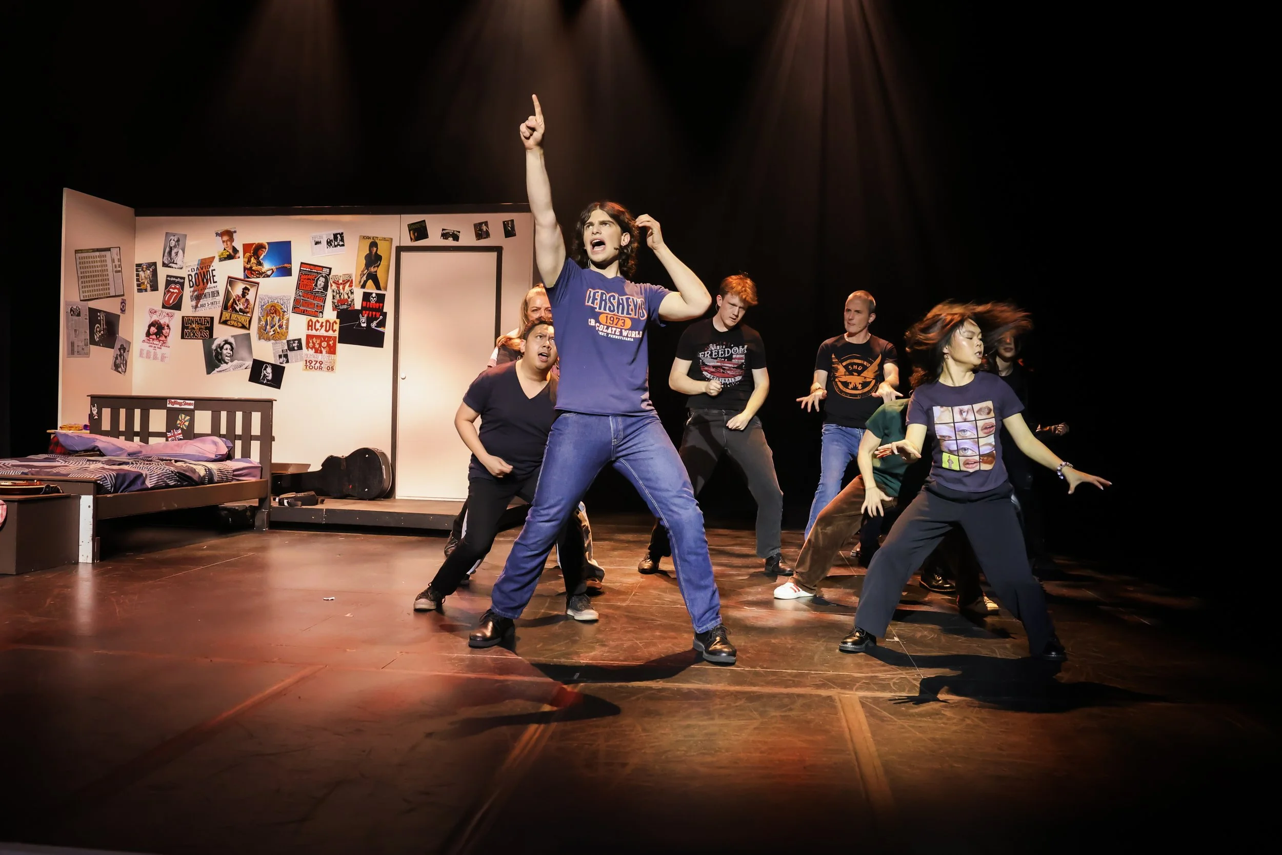 Group of young actors rehearsing a play on stage, with one central figure pointing upward and others dancing, set in a bedroom with posters and photos on the wall.