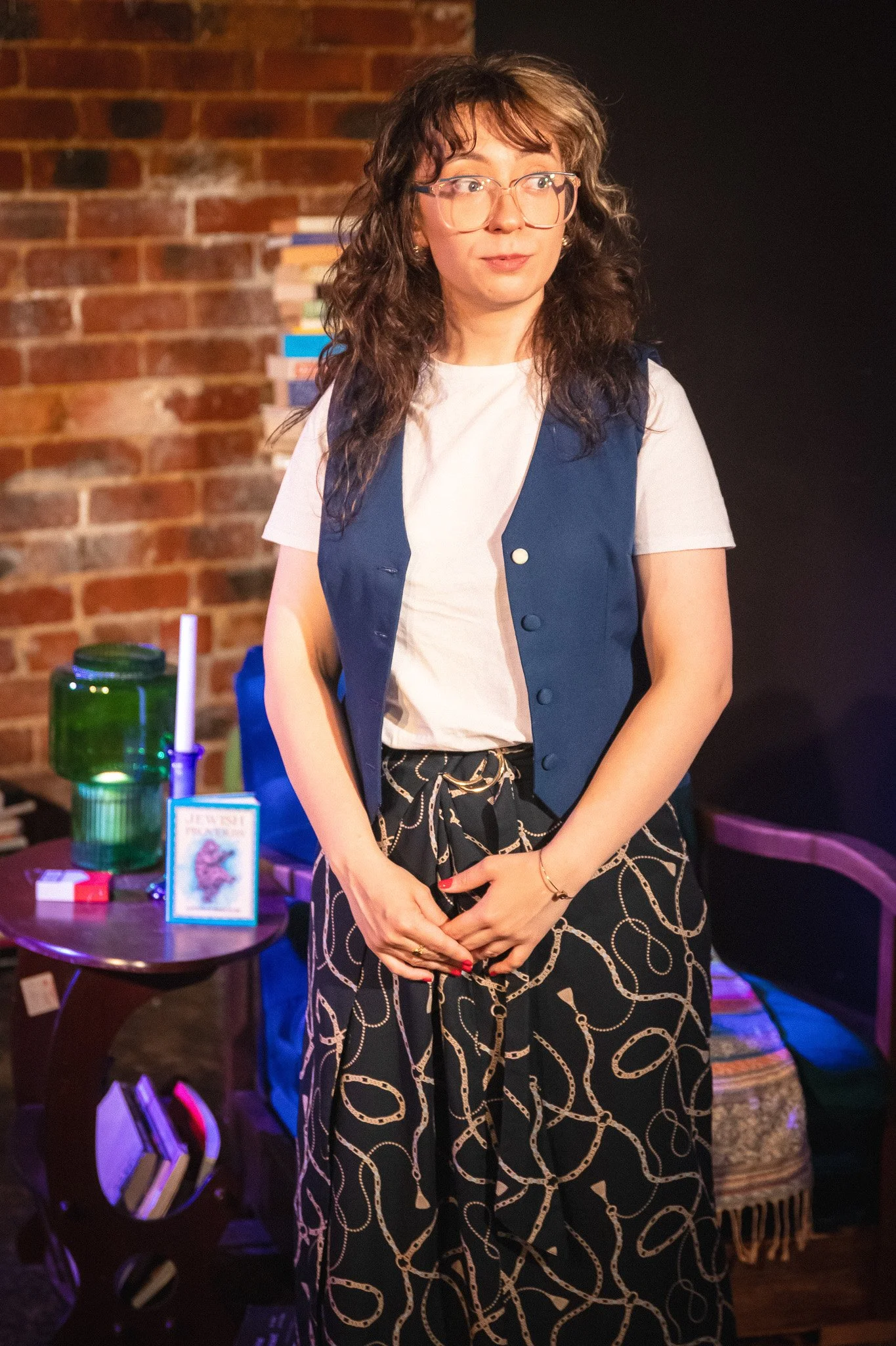 A woman with curly hair and glasses standing near a table with books, a green jar, and a lit candle in a green holder, in front of a brick wall.