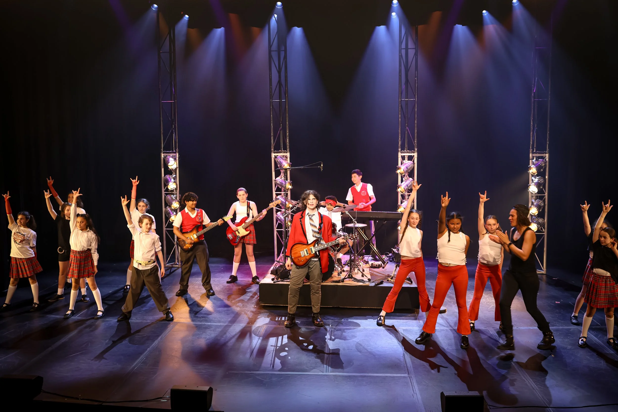 Children performing on stage with musical instruments, under colorful stage lights, some dancing and others playing instruments, in a school or theatrical setting.