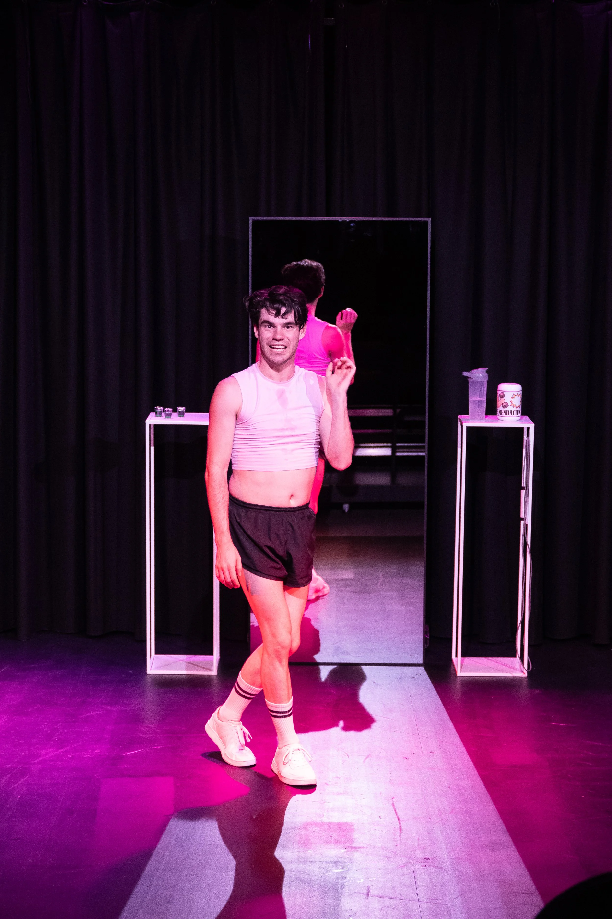 A performer dressed in a white tank top, black shorts, and white sneakers stands on stage in front of a mirror, with pink stage lighting and black curtains in the background.