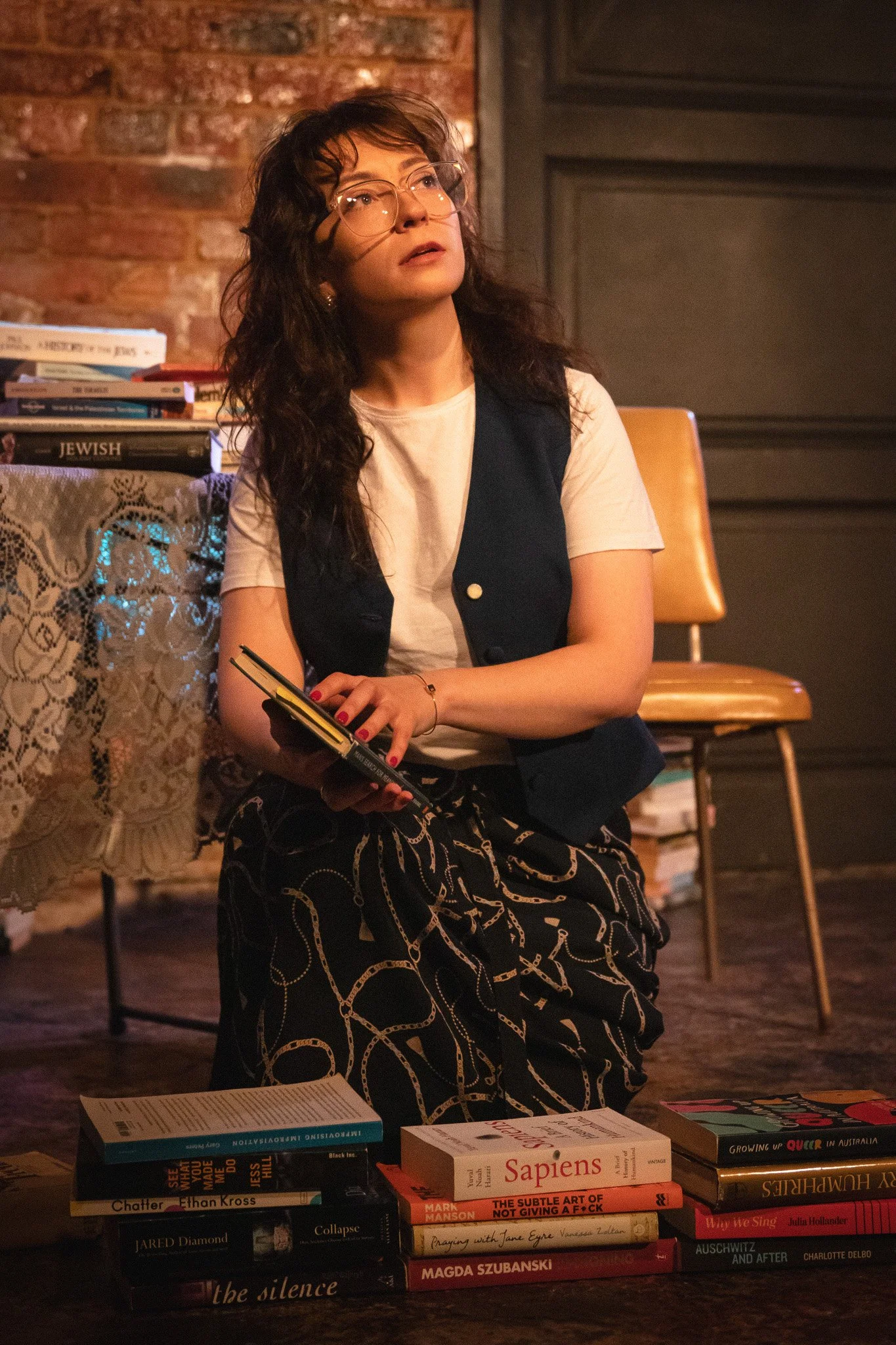 A woman sitting on the floor in a room with exposed brick wall, holding a book and surrounded by stacks of books, with glasses and a thoughtful expression.