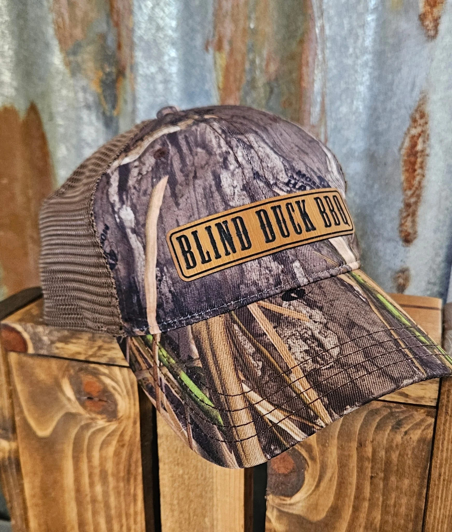 Close-up of a camouflage baseball cap with a leather patch on the front reading Blind Duck BBQ.
