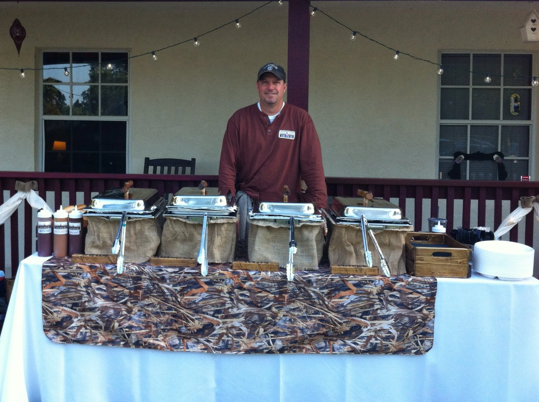 Pit-Matt-ster, Matt Morgan, behind the serving table at a wedding catering event with buffet trays, bottles of sauces, and condiments on a decorated table, outdoors on a porch or patio with string lights and windows in the background.