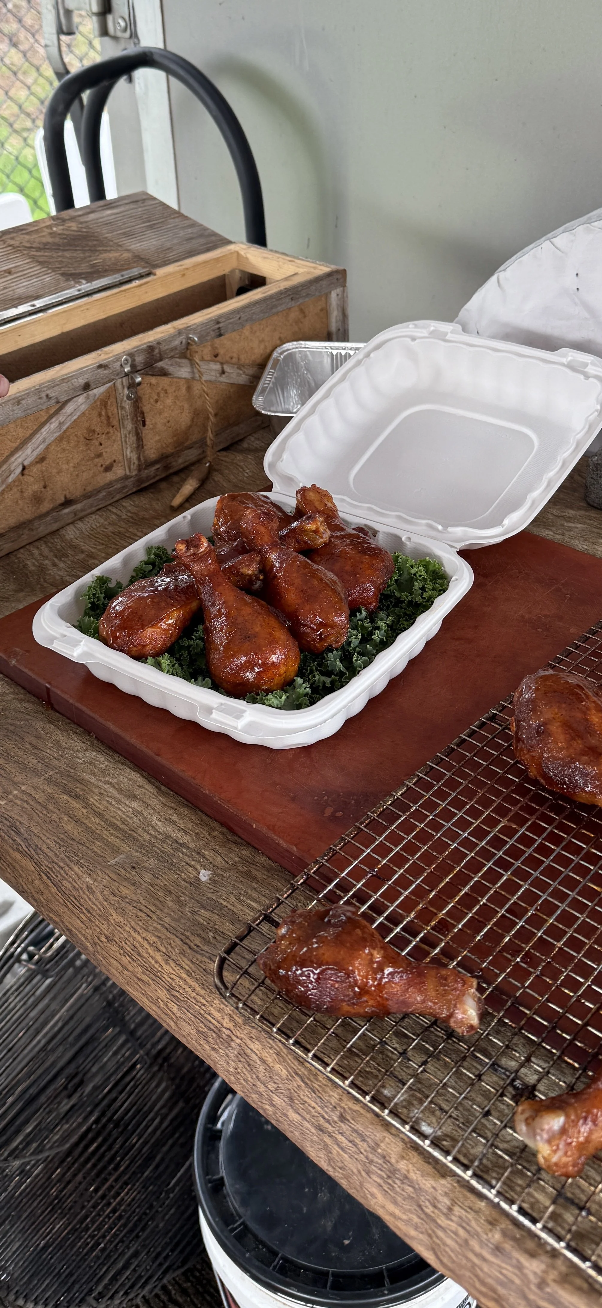 Container of glazed chicken drumsticks on a bed of greens, with additional drumsticks and pieces on a wire rack nearby.