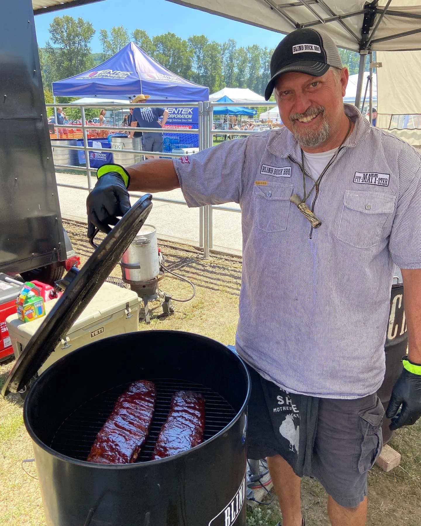 Pit-Matt-ster, Matt Morgan, grilling two racks of barbecue ribs in a barrel smoker at an outdoor BBQ competition event, with tents and people in the background.