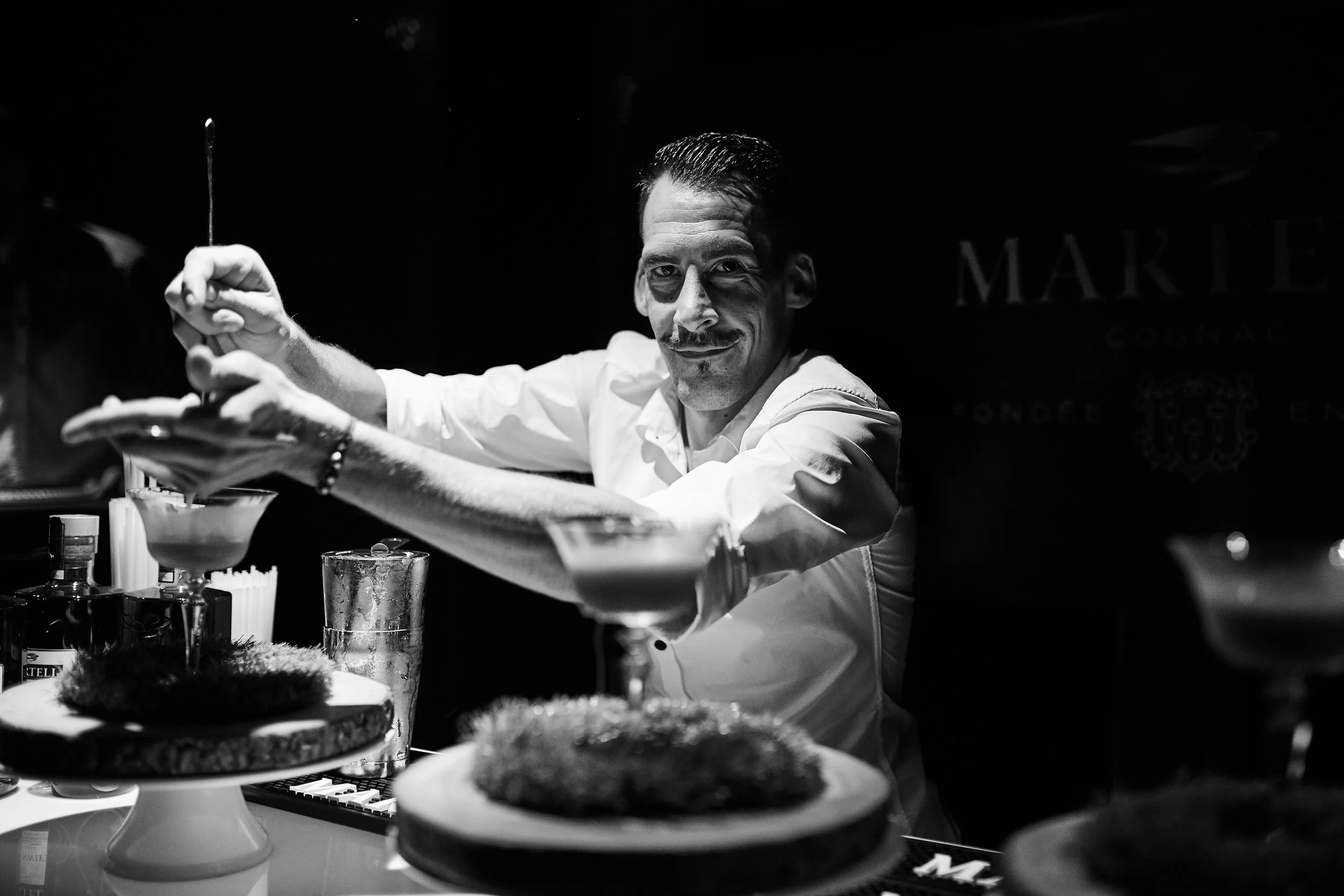 A bartender preparing a cocktail behind a bar with glasses and bottles, smiling, in black and white.