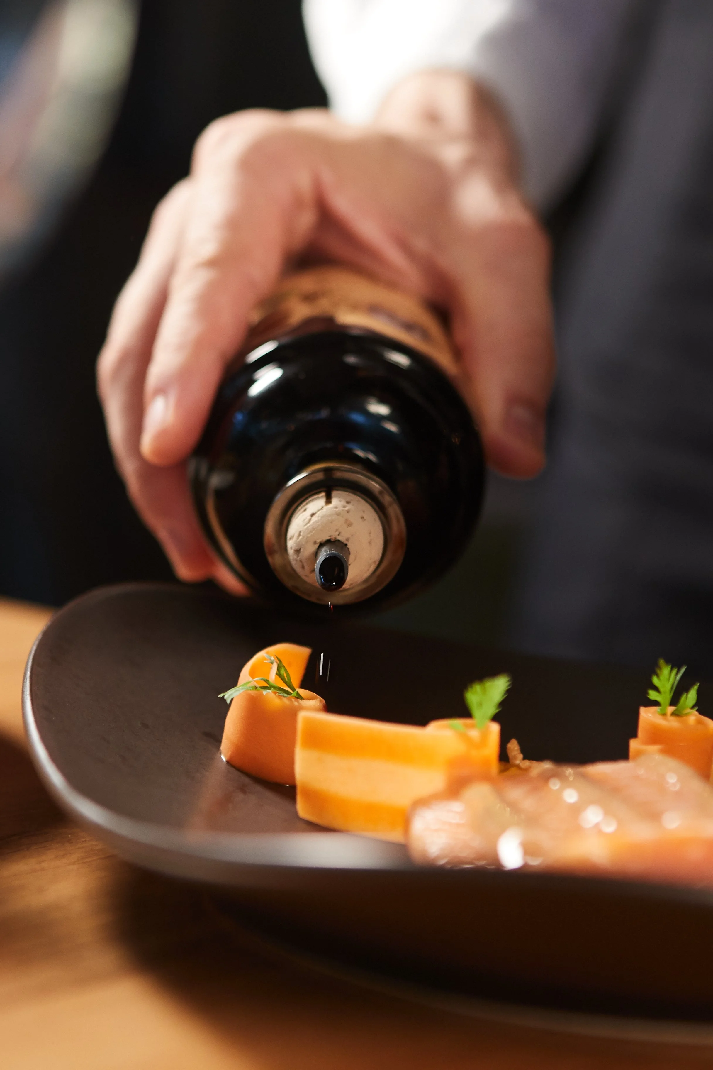 A person holding a dark bottle upside down over a black plate with sliced cured meat garnished with small green herbs, likely being seasoned or prepared for serving.