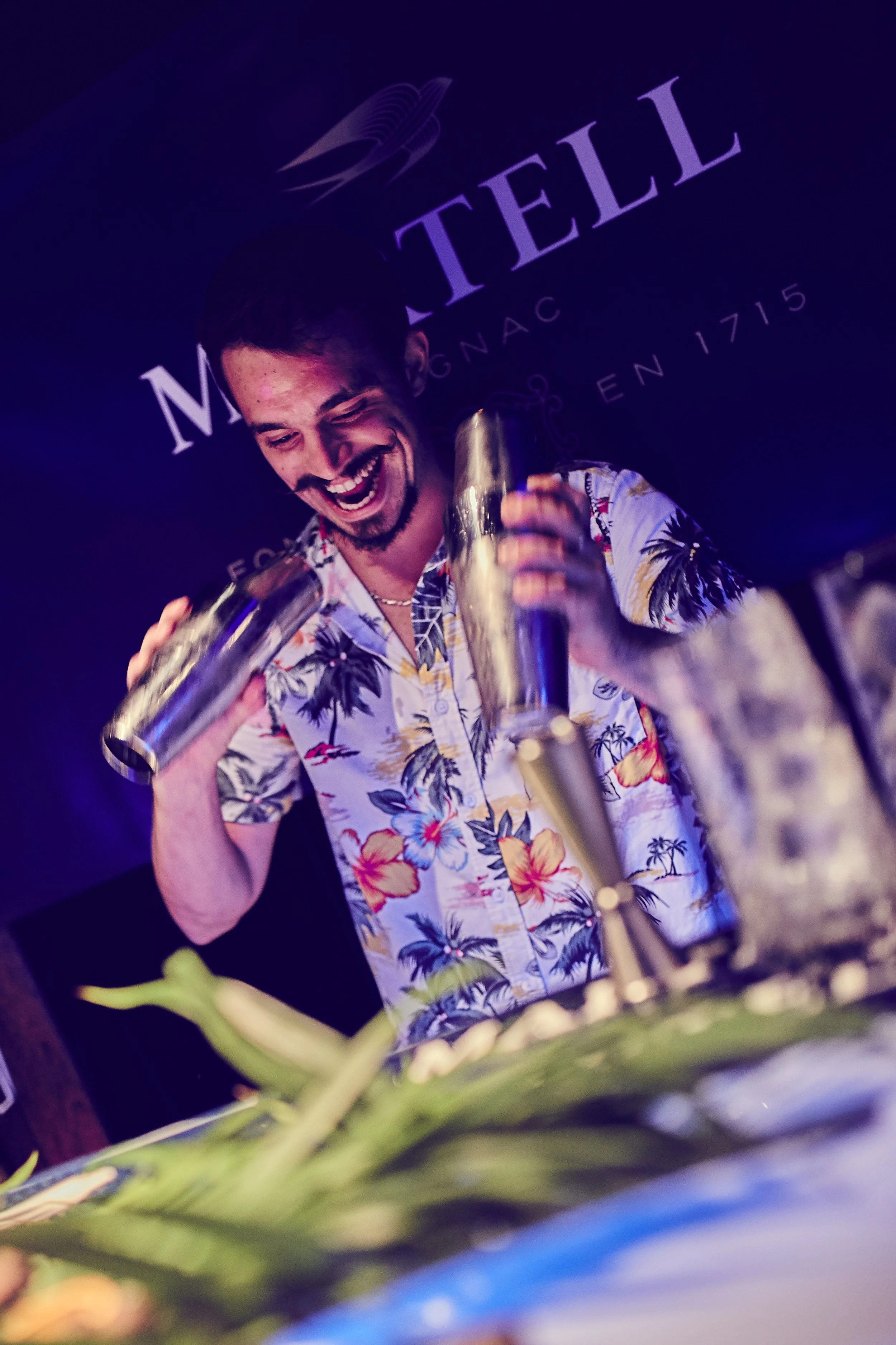 A man wearing a colorful Hawaiian shirt is smiling and laughing while holding a cocktail shaker at a bar. There are glasses and a plant in the foreground. A sign behind him reads 'MOTELL' with additional text.