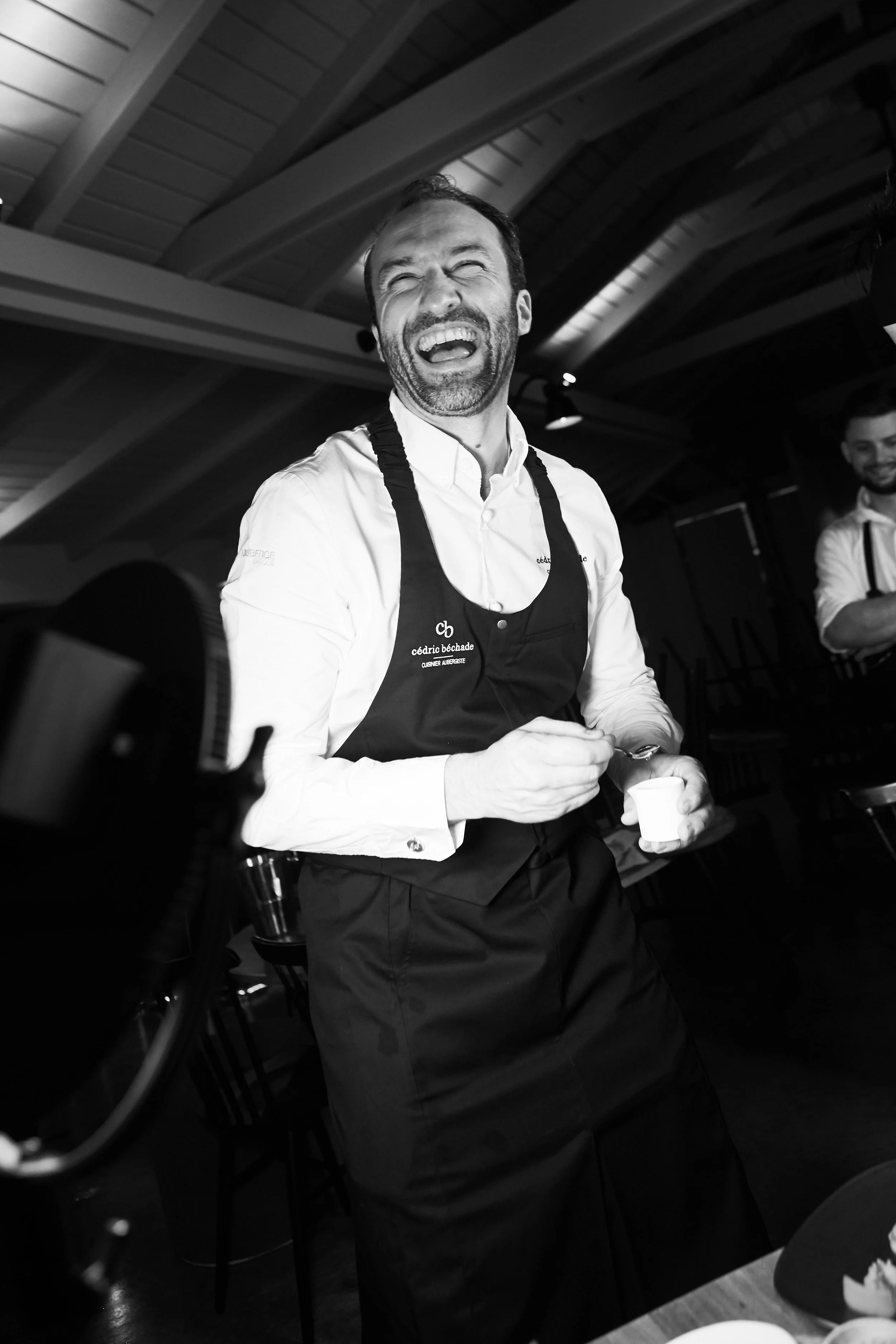 A man with a beard laughing, holding a small container and a spoon, wearing a white shirt and black apron, in a restaurant or kitchen setting.
