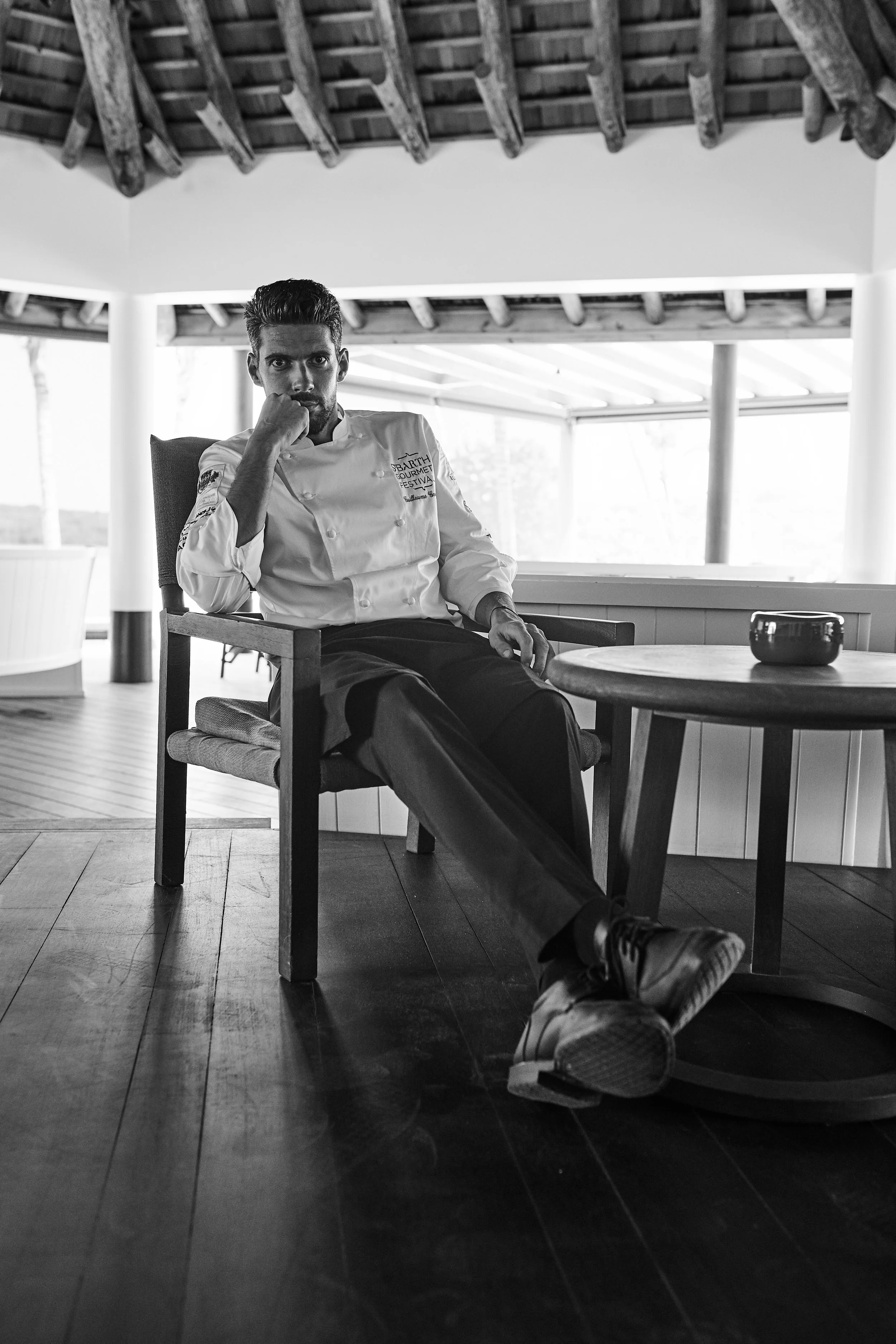 A man in a chef's uniform sitting in a chair indoors, looking at the camera with a serious expression, with a round table in front of him and a small bowl on it, in a room with wooden ceiling beams and large windows.