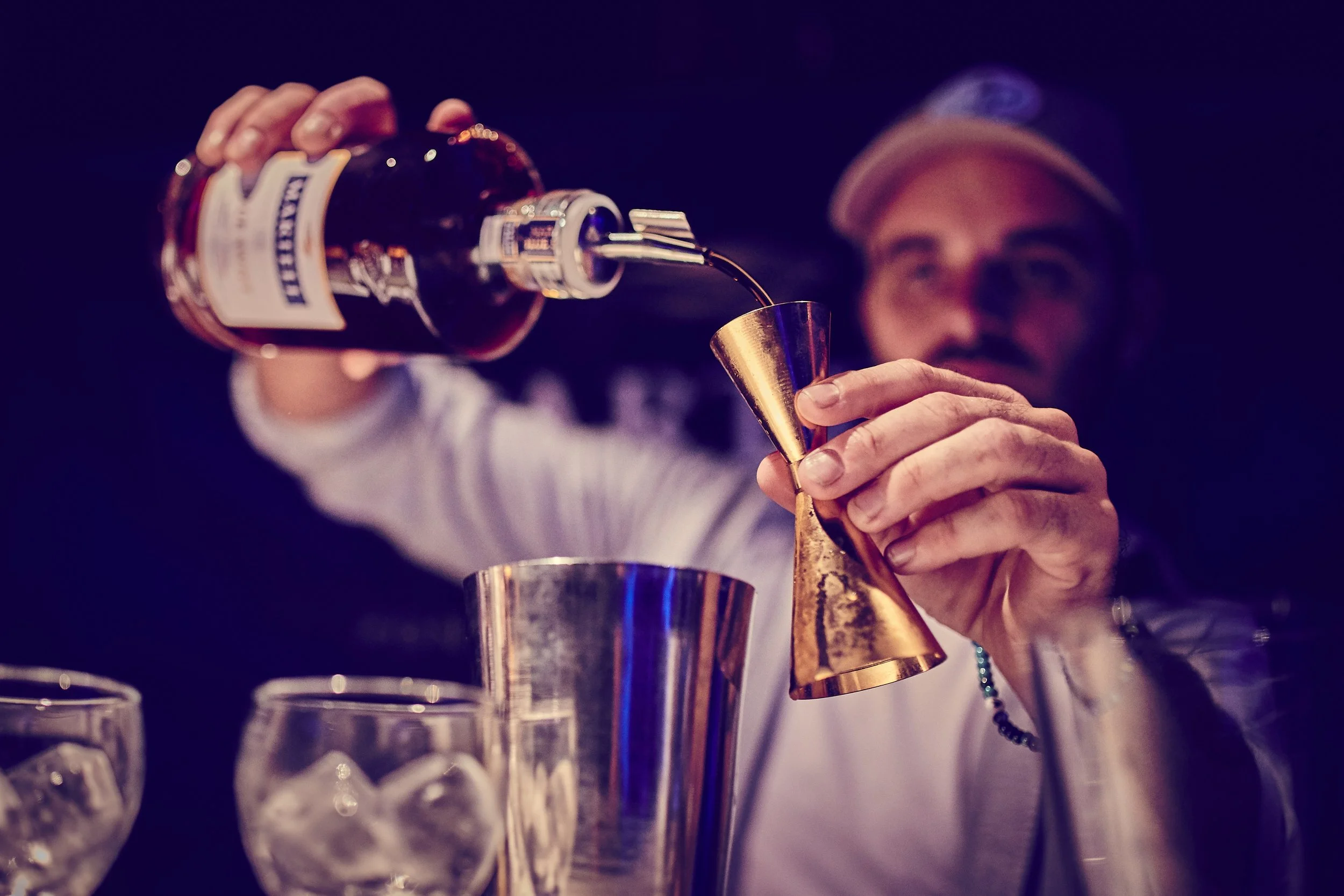 A man wearing a cap pours a dark alcoholic drink from a bottle into a gold-colored jigger, with a dark background and glasses of ice in the foreground.