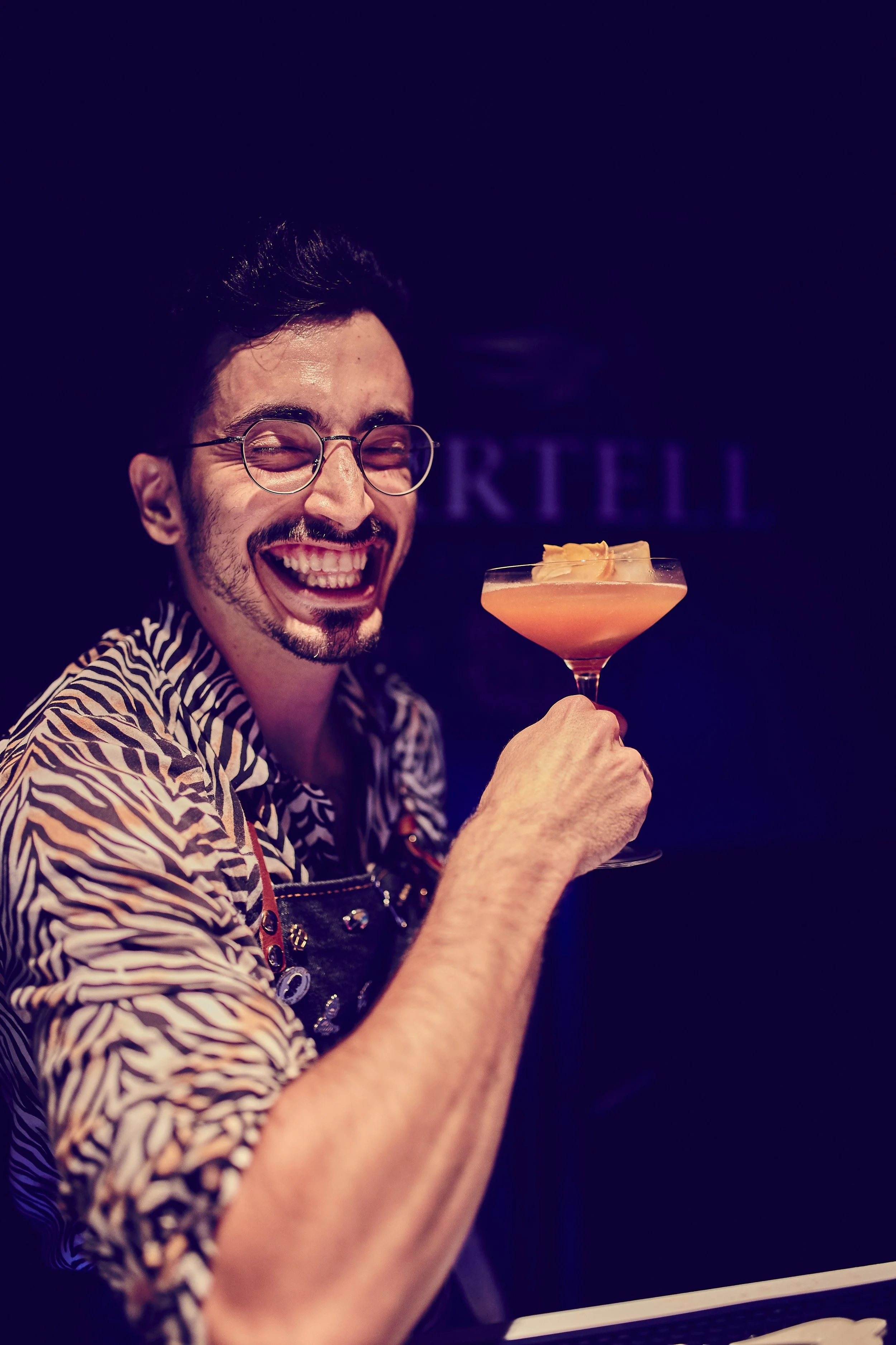 A smiling man with glasses holding a pink cocktail in a martini glass.