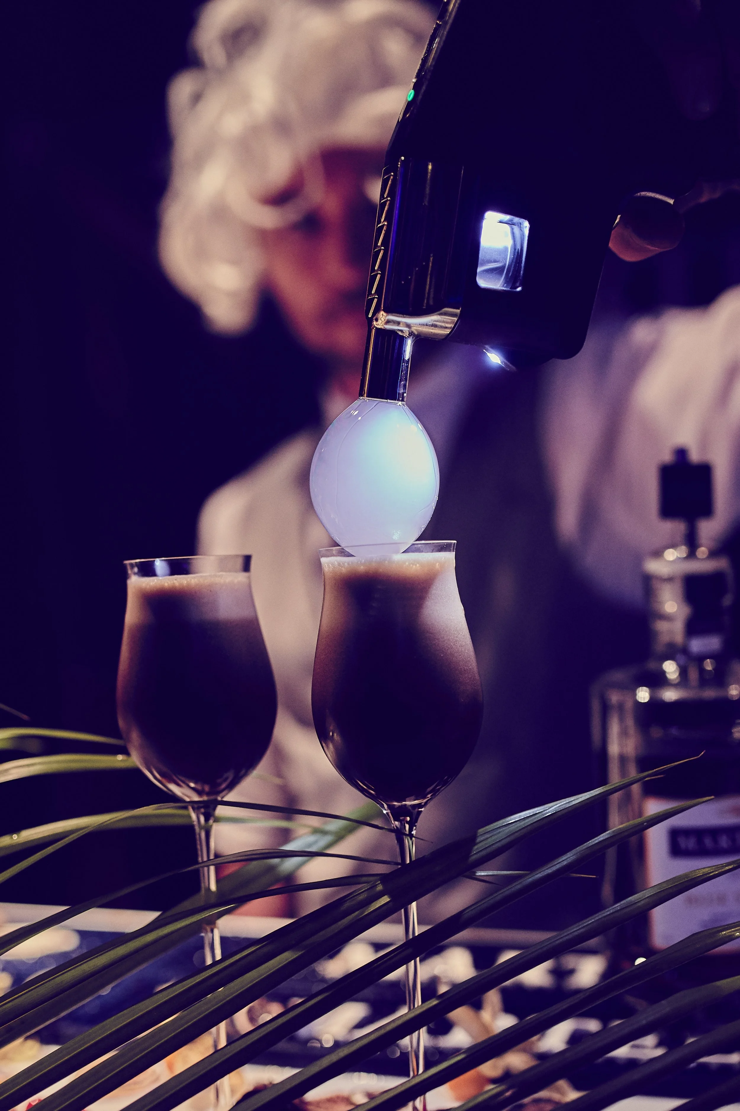 A person with curly gray hair pouring a drink with a ladle from a siphon into two cocktail glasses, with a blurred background and decorative leaves in the foreground.