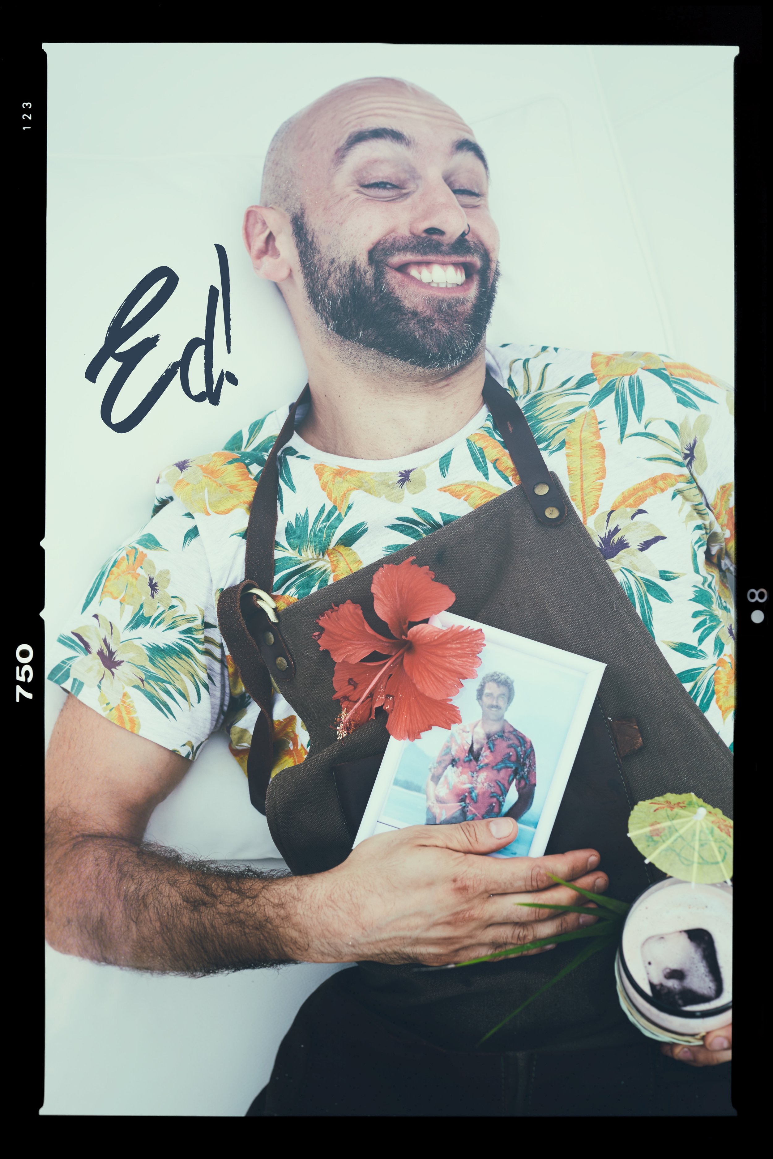 A man with a beard and a bald head smiling and lying down, holding a photo, a cocktail, and wearing a colorful tropical shirt and an apron. The image has tropical decorations like a hibiscus flower and a small citrus umbrella.