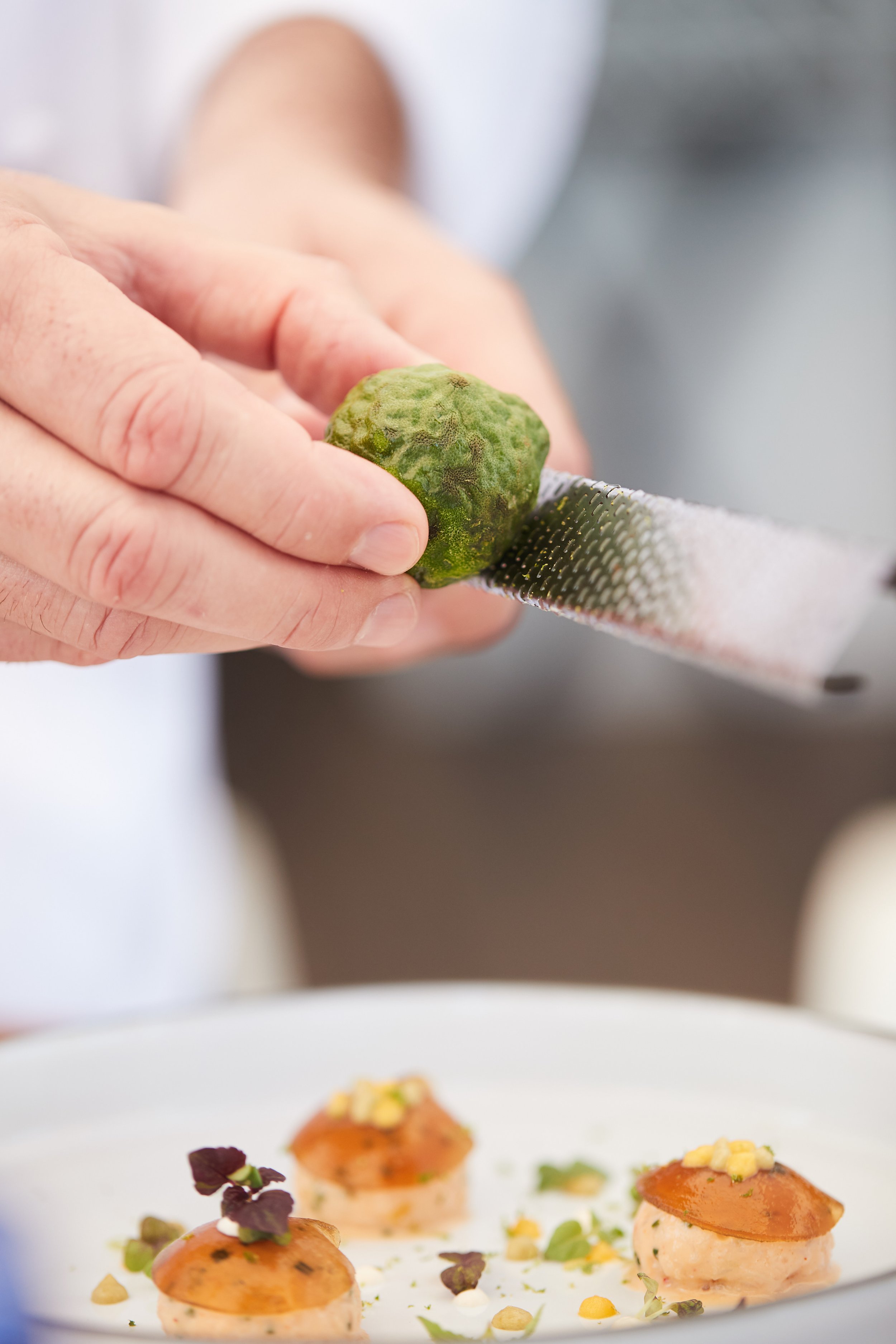 Close-up of a person grating a green citrus fruit over a plate with small appetizers garnished with herbs and edible flowers.
