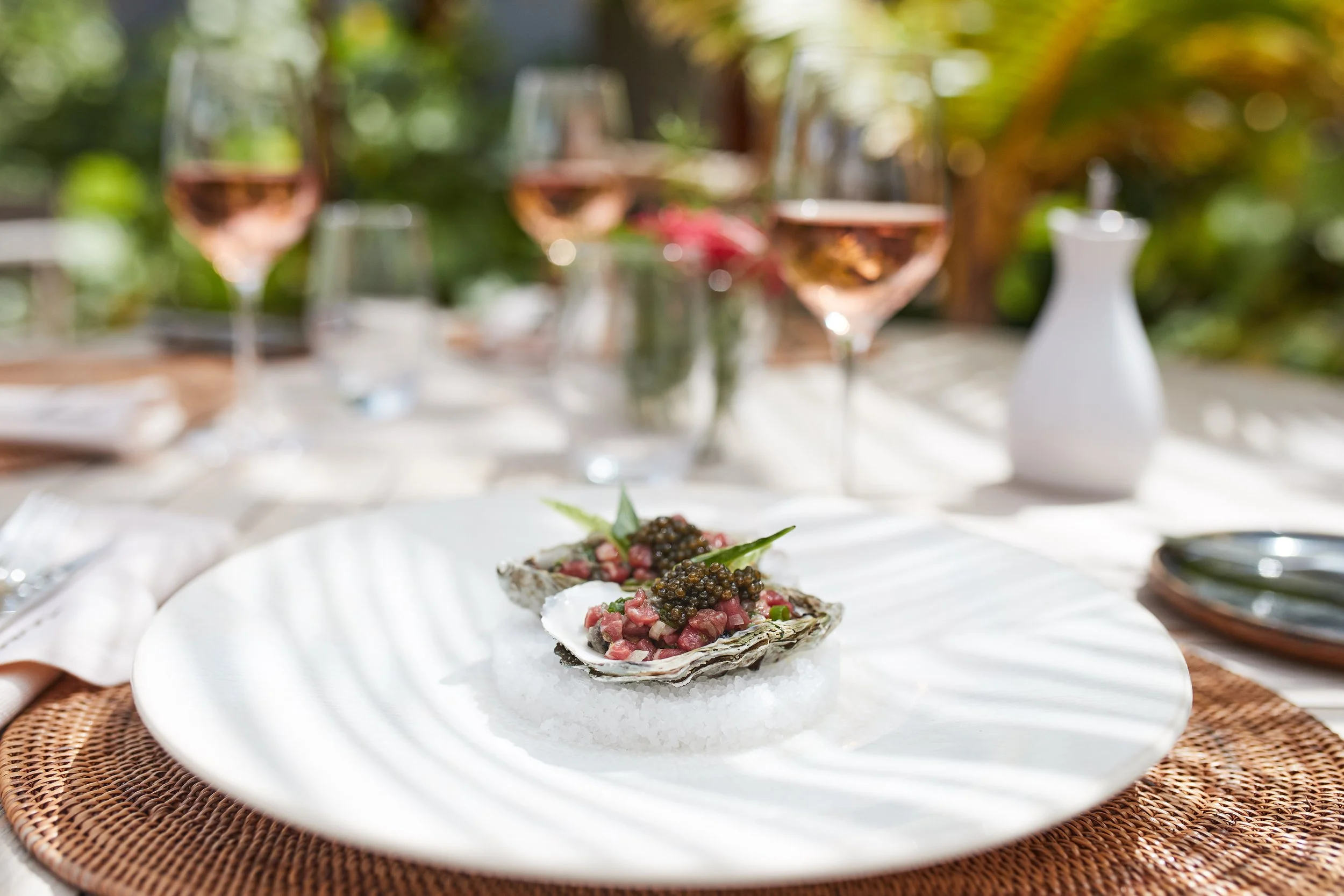 Oysters topped with caviar and garnishes on a white plate with glasses of rosé wine and a white vase in the background.