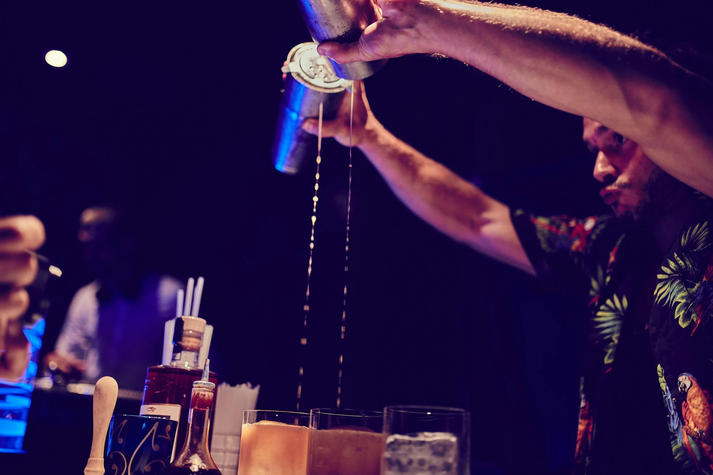 A bartender pouring liquid from a shaker into a glass at a bar with various bottles and tools on the counter, while people are in the background.