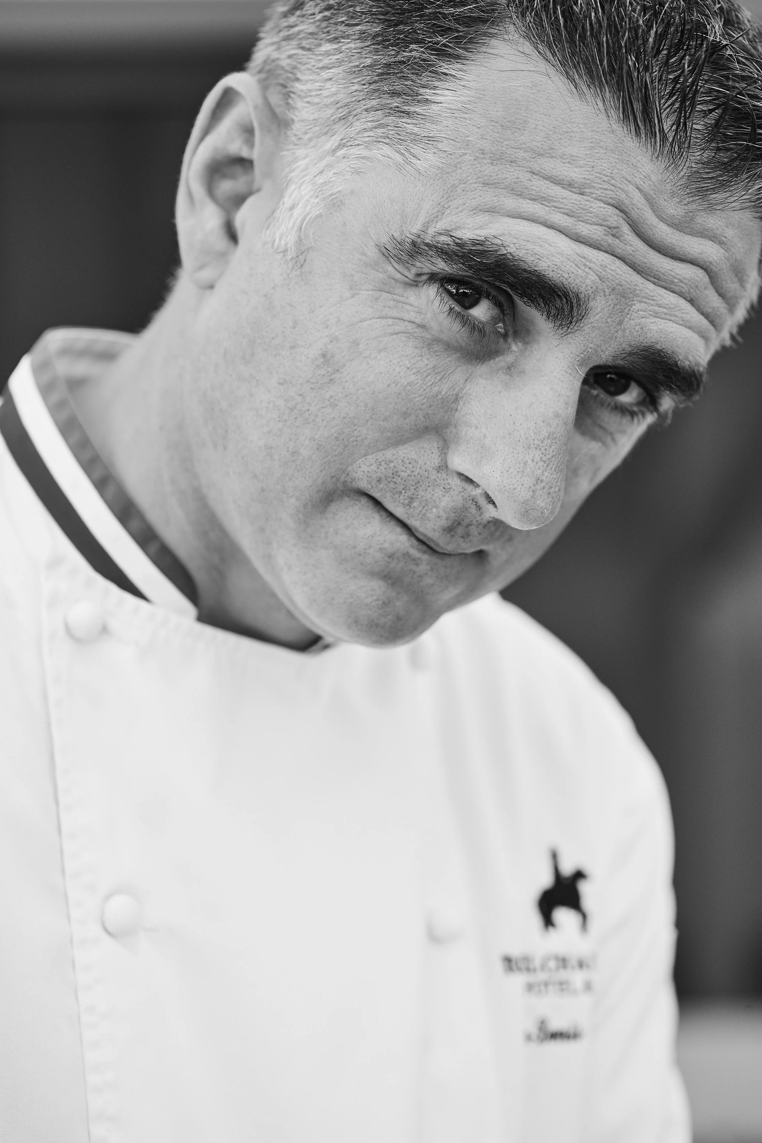 Close-up black and white photograph of a male chef in a white uniform, with a serious expression, looking directly into the camera.