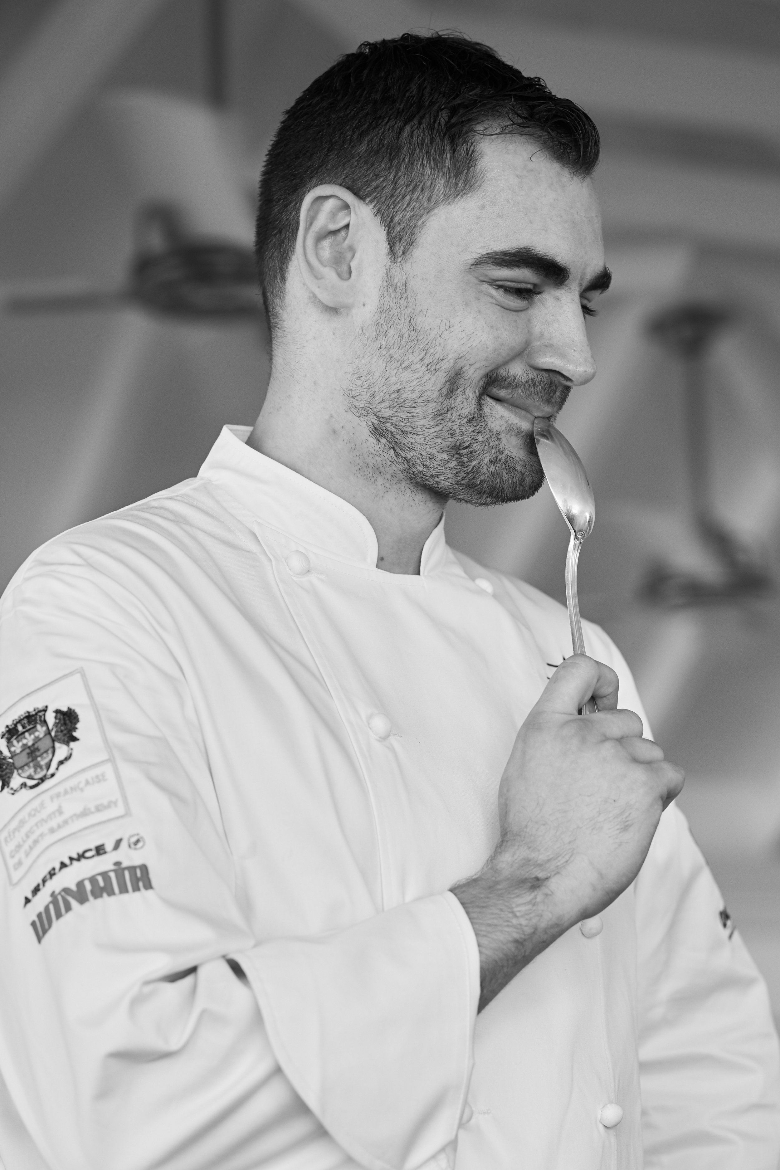 A smiling male chef in a white uniform tasting food with a spoon in a professional kitchen.