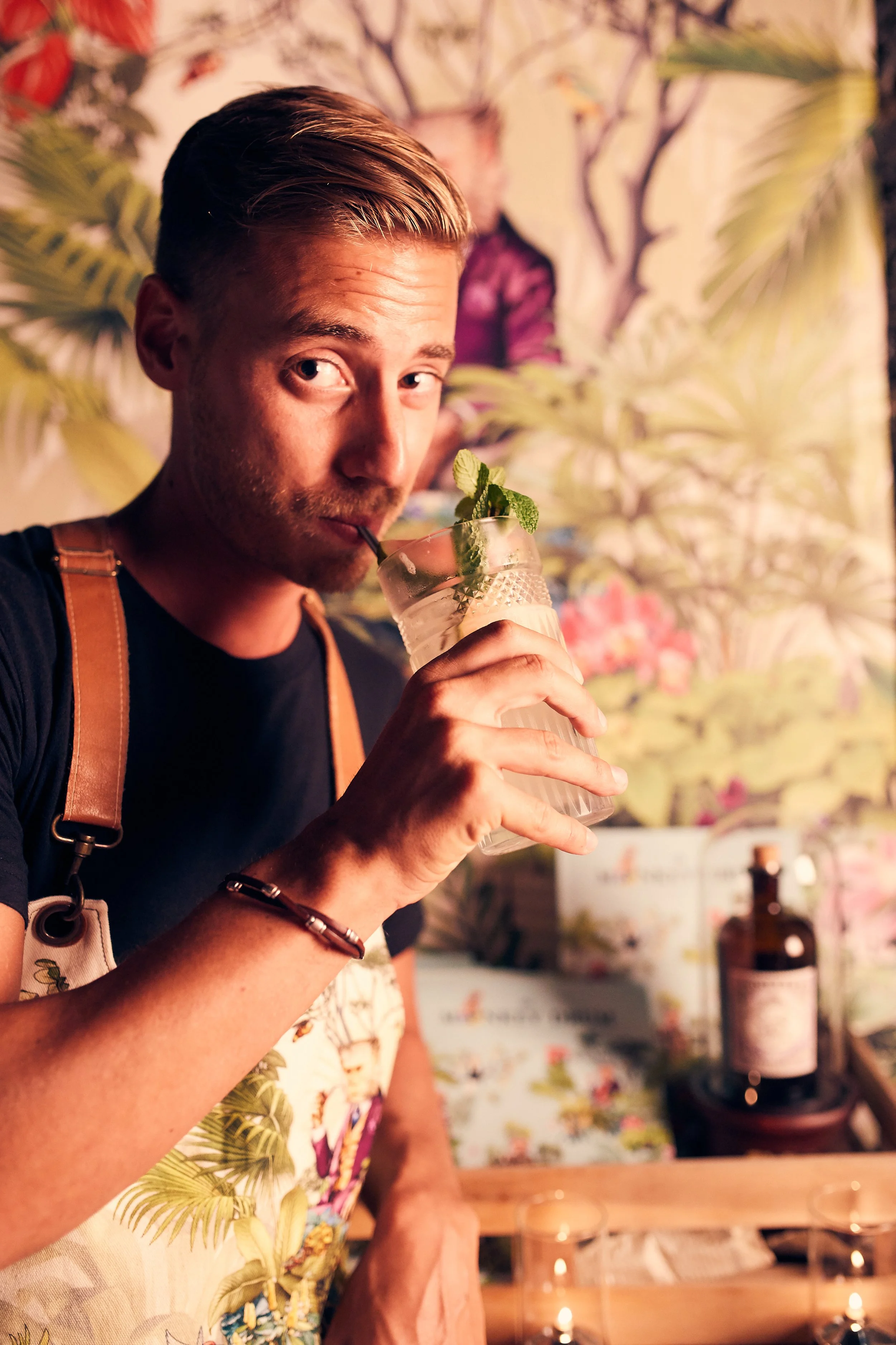 A man with blond hair and a black shirt drinks a cocktail with a straw, garnished with mint leaves, in a dimly lit setting with tropical-themed decor and bottles in the background.