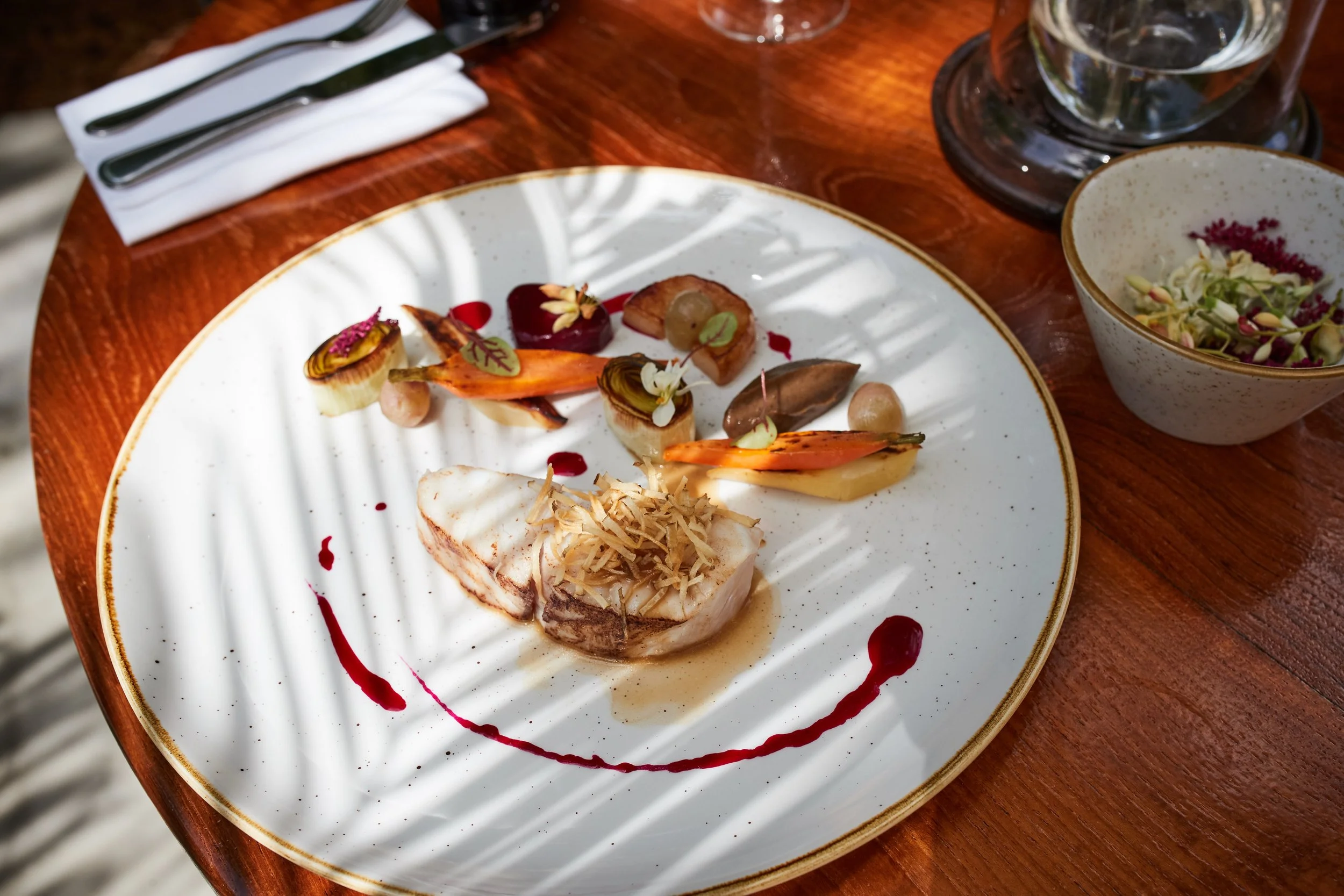 A gourmet dish featuring a cooked fish fillet garnished with shredded vegetables, served with assorted vegetables, herbs, and sauces on a white plate with a gold rim, placed on a wooden table with wine glasses and a bowl of salad nearby.