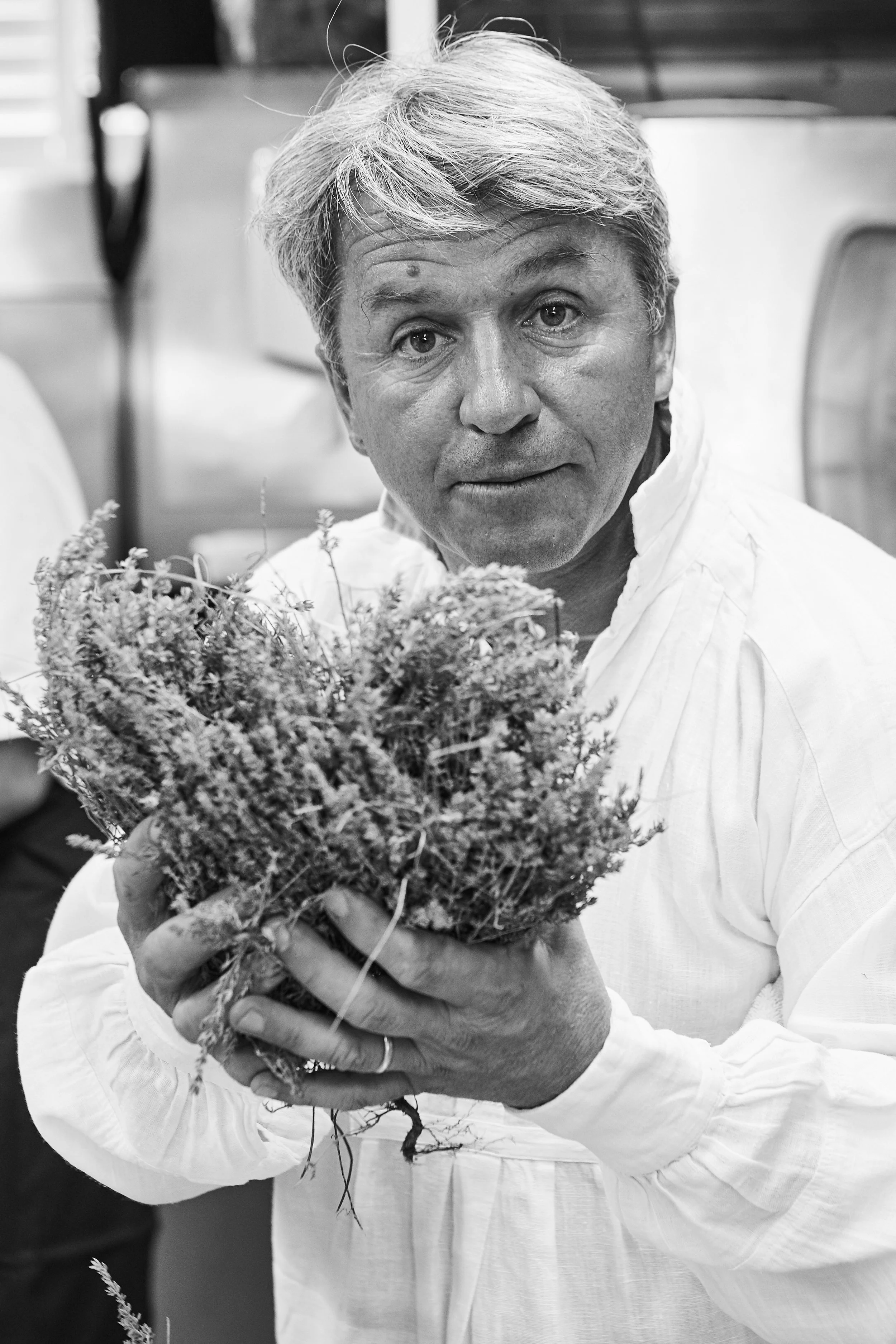 A man with light-colored hair wearing a white shirt holds a bunch of flowers or herbs and looks at the camera. The photo is in black and white.