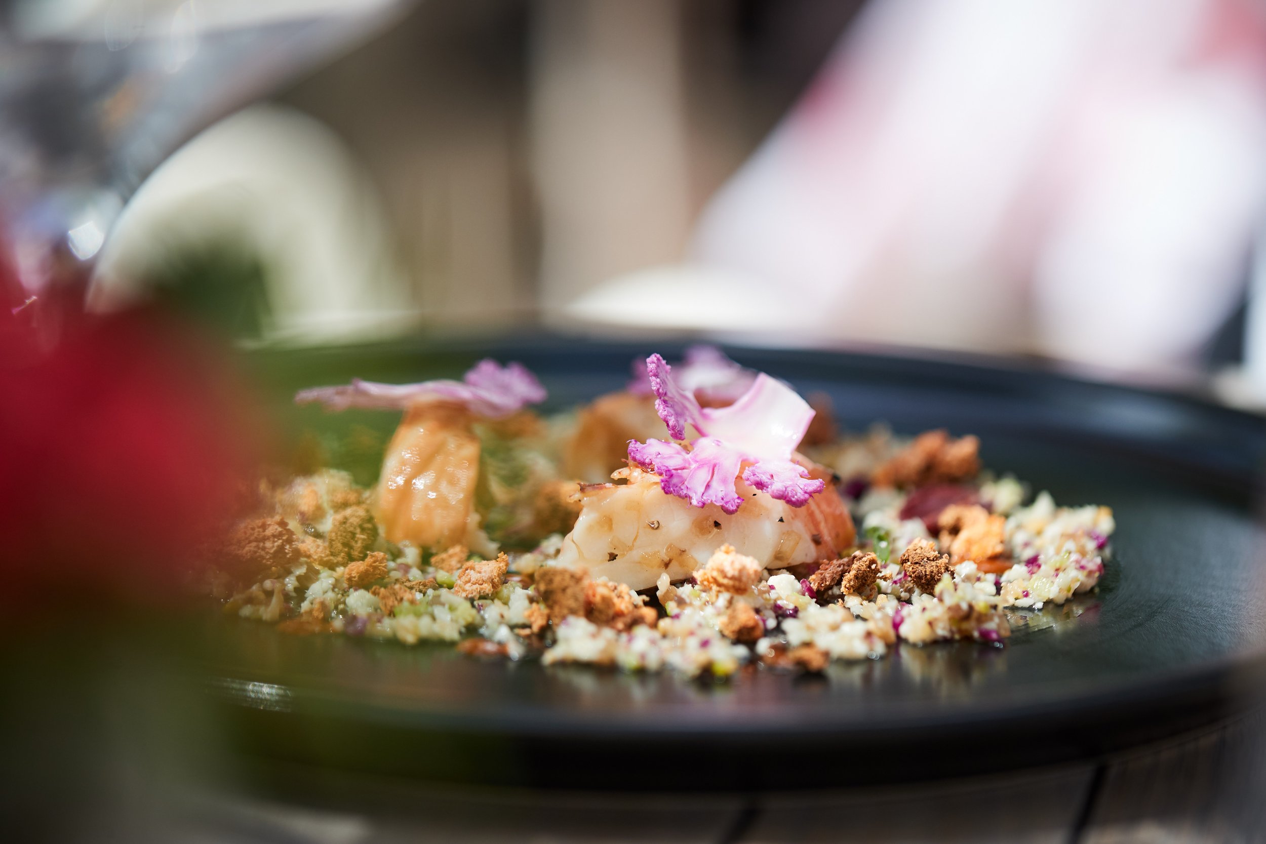 Close-up of a gourmet dish on a black plate, featuring a piece of seafood topped with pink edible flower petals, surrounded by various textured ingredients and garnishes.