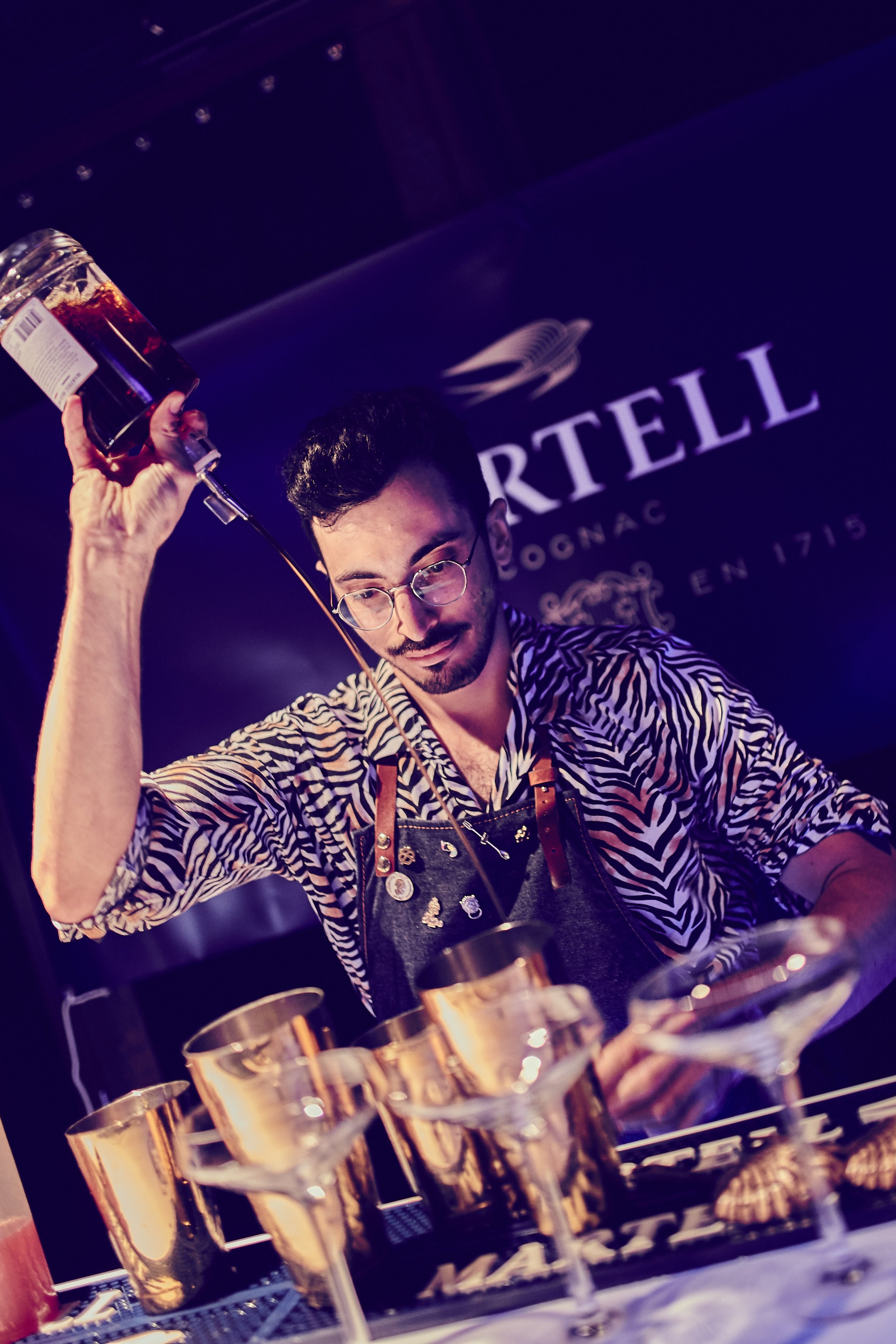 Bartender pouring a drink into a cocktail glass at a bar, with a sign in the background.