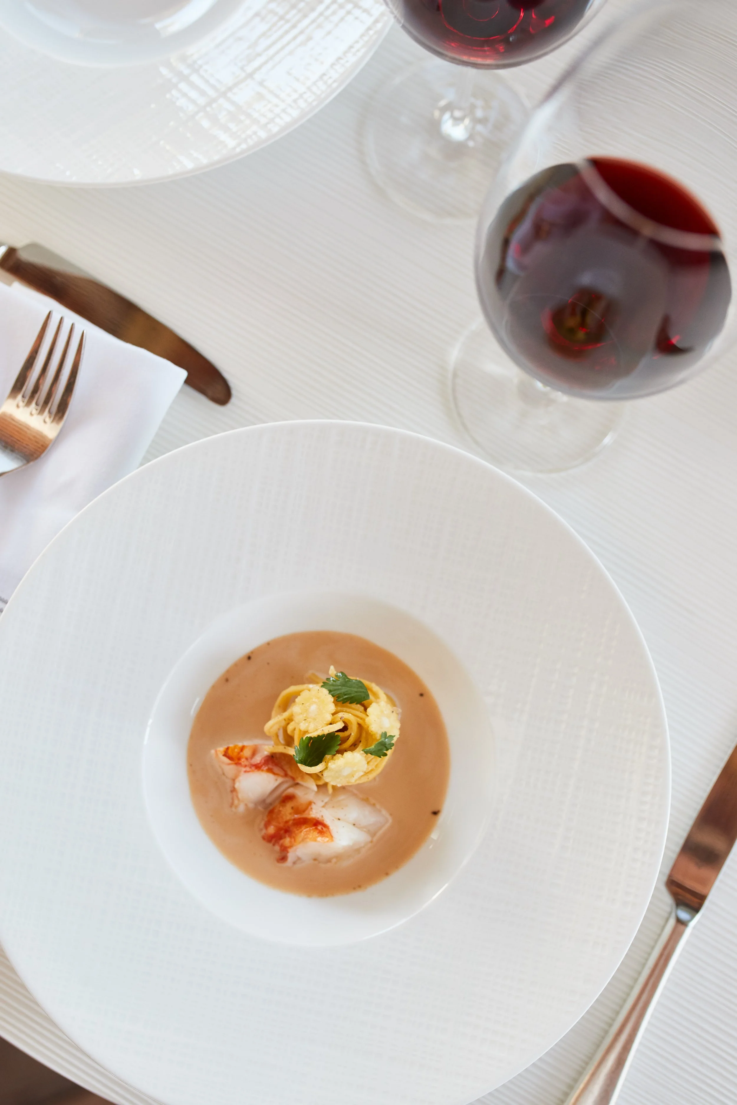 A fancy dining table setting with a bowl of lobster bisque garnished with fresh herbs and a pasta nest on top, two glasses of red wine, a white napkin, and a silver fork and knife.