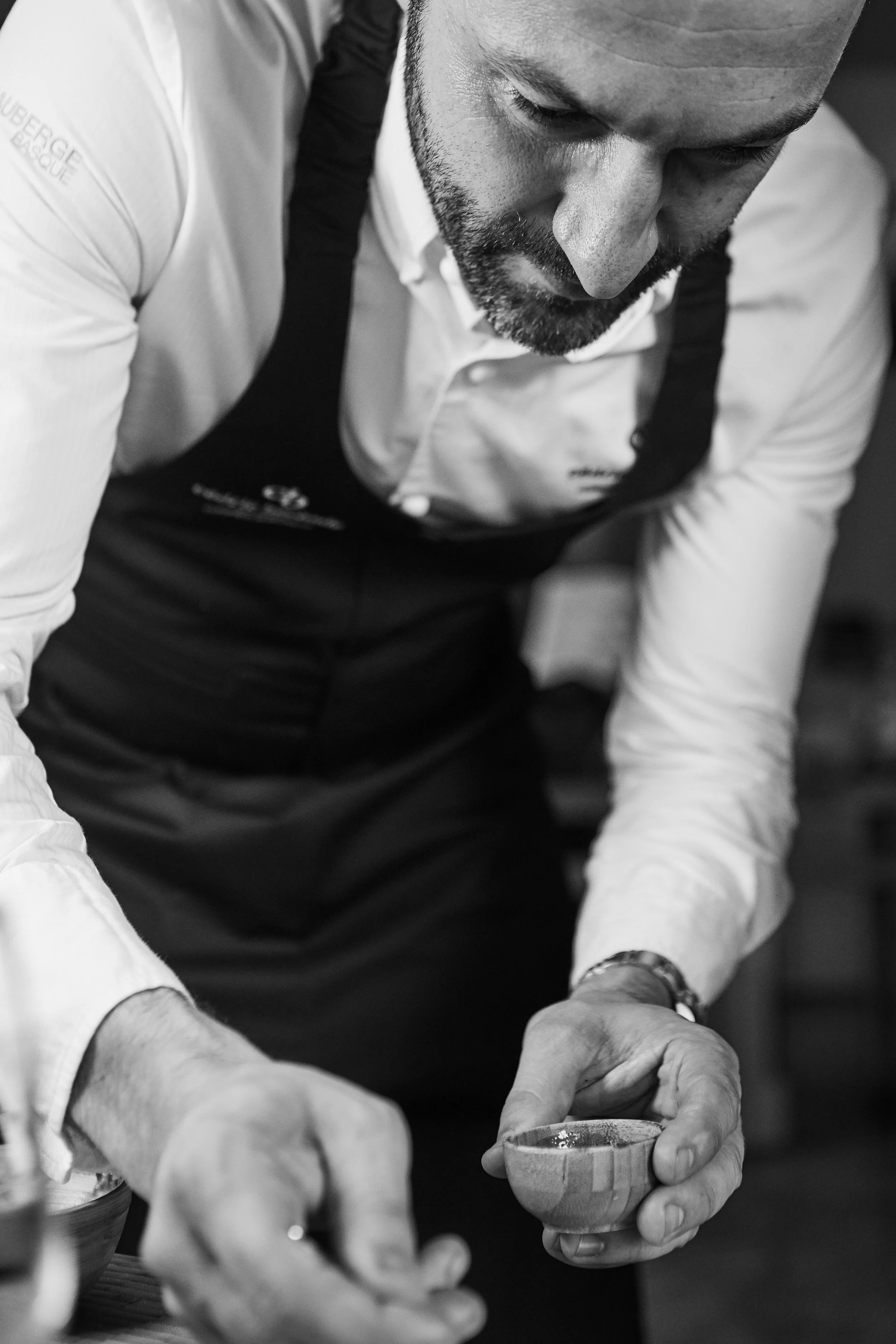 A man with a beard dressed in a white shirt and apron, leaning over a table, holding a small wooden bowl.