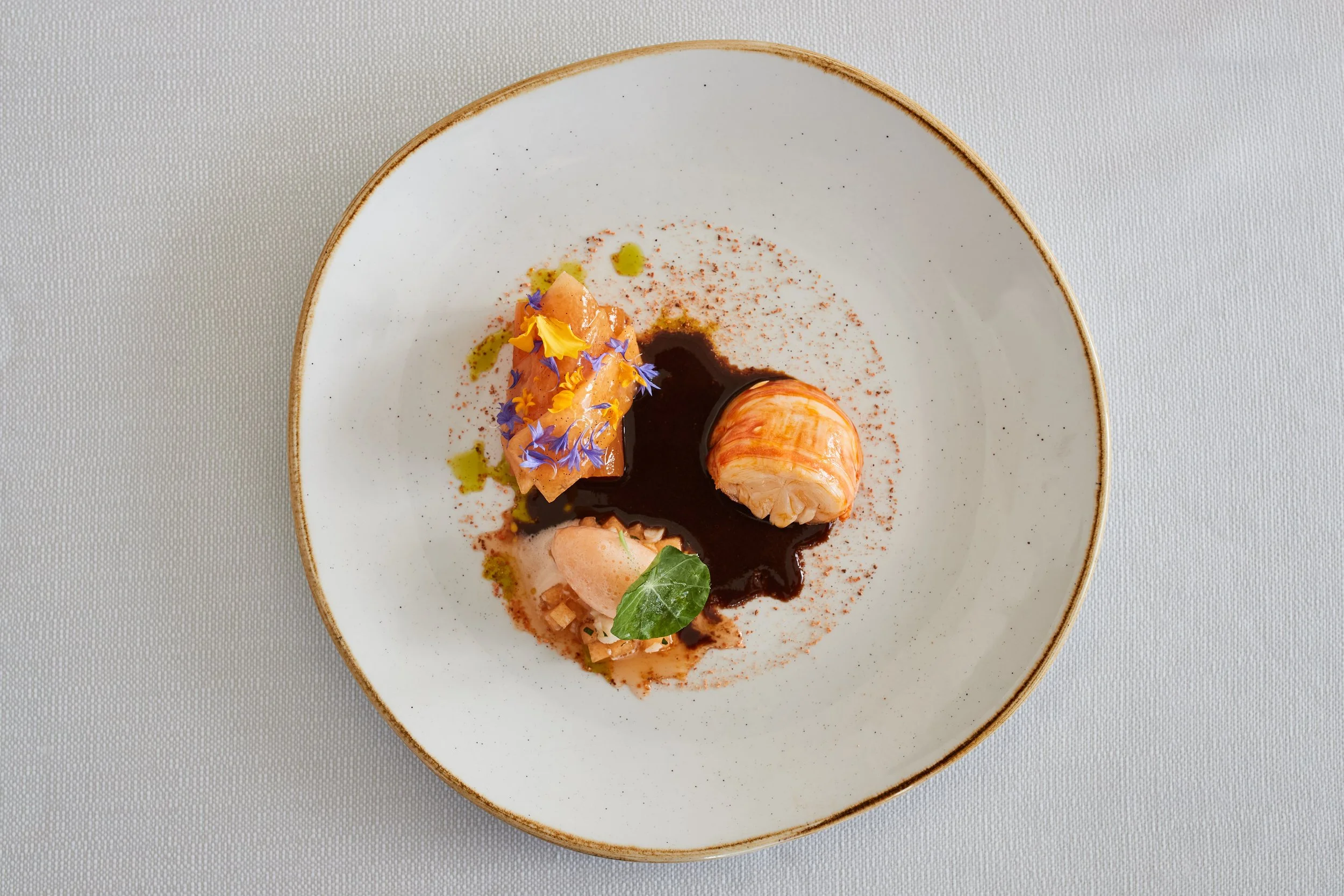 A gourmet dish with three pieces of seafood on a white plate, garnished with edible flowers, herbs, and sauces, served with artistic presentation.