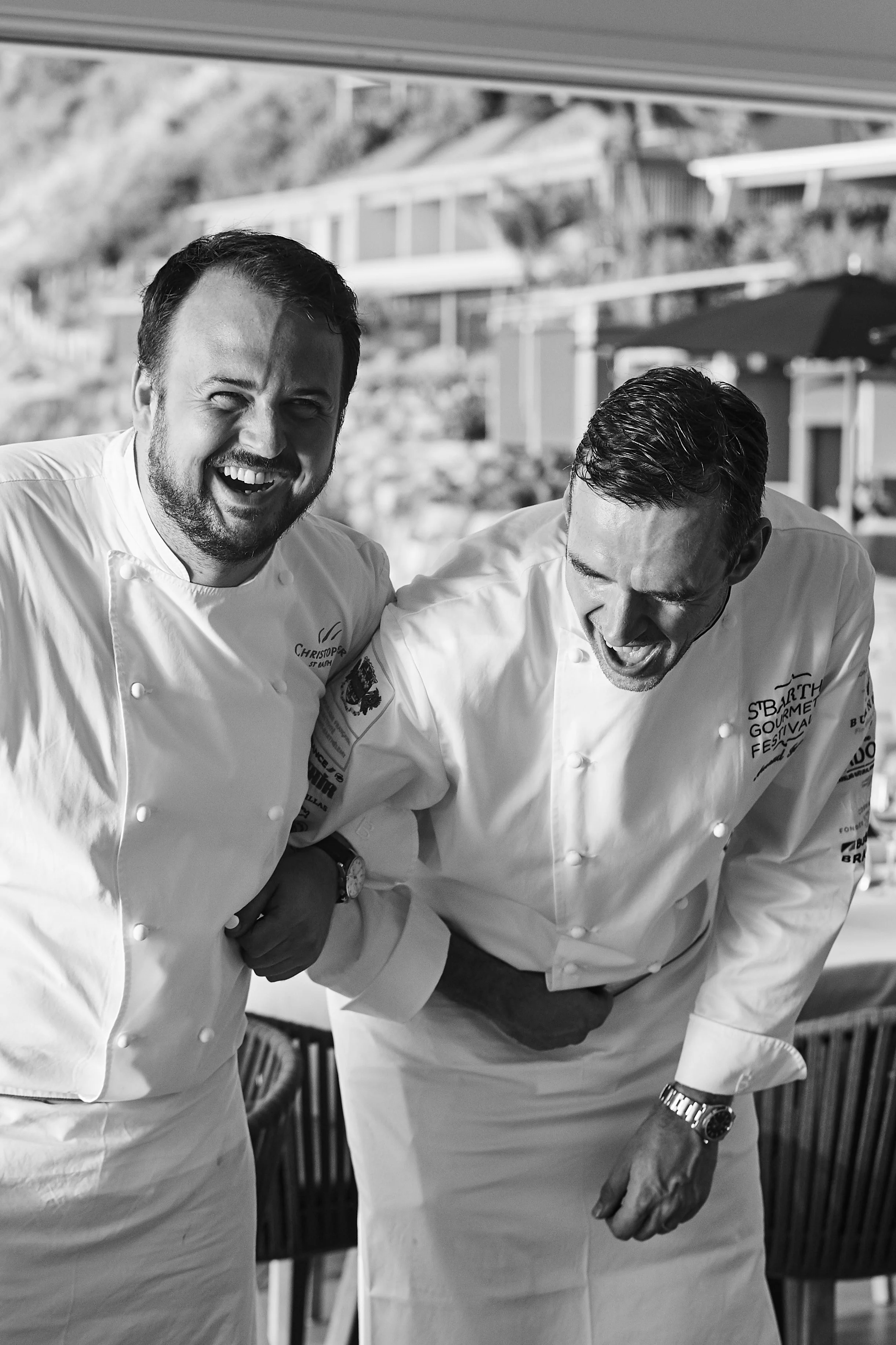 Two male chefs in white uniforms laughing and playfully holding each other's arms.