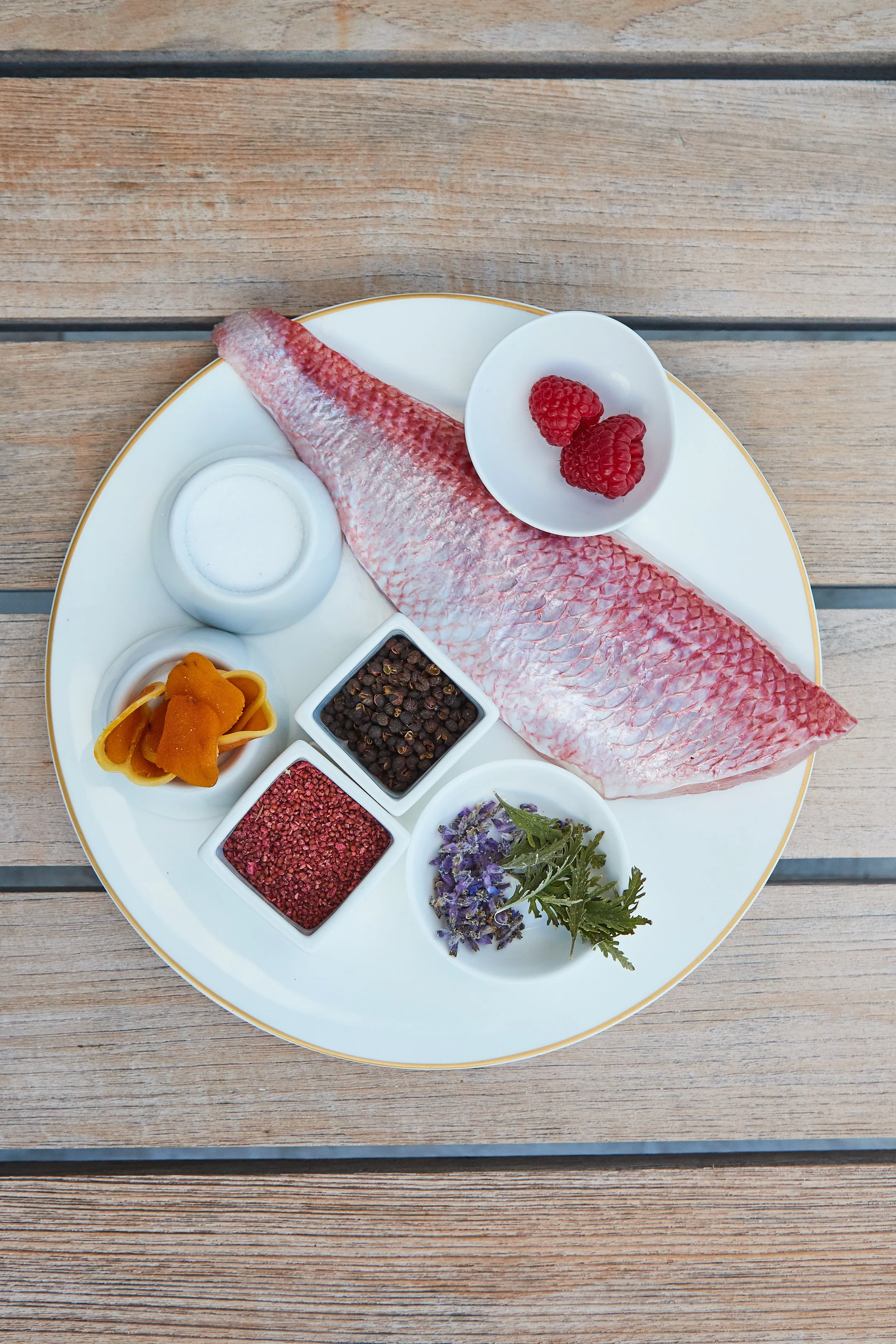 Fresh raw fish fillet on a white plate with spices, herbs, and berries, placed on a wooden surface.