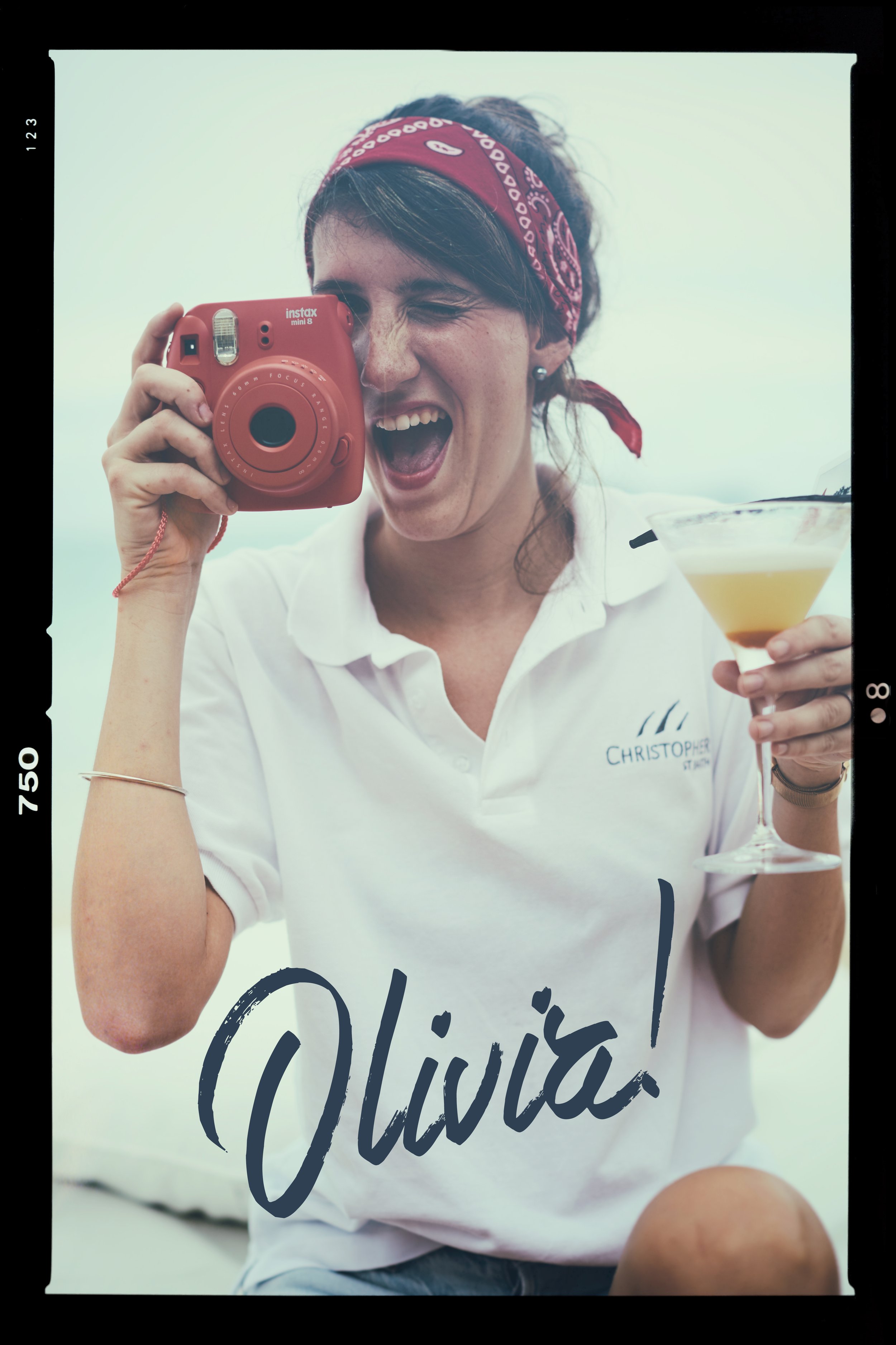 A woman wearing a red bandana and white polo shirt with 'Christopher' embroidered on it, holding a red instant camera up to her face and a cocktail glass in her other hand, with the name 'Olivia!' written in large script on the photo.