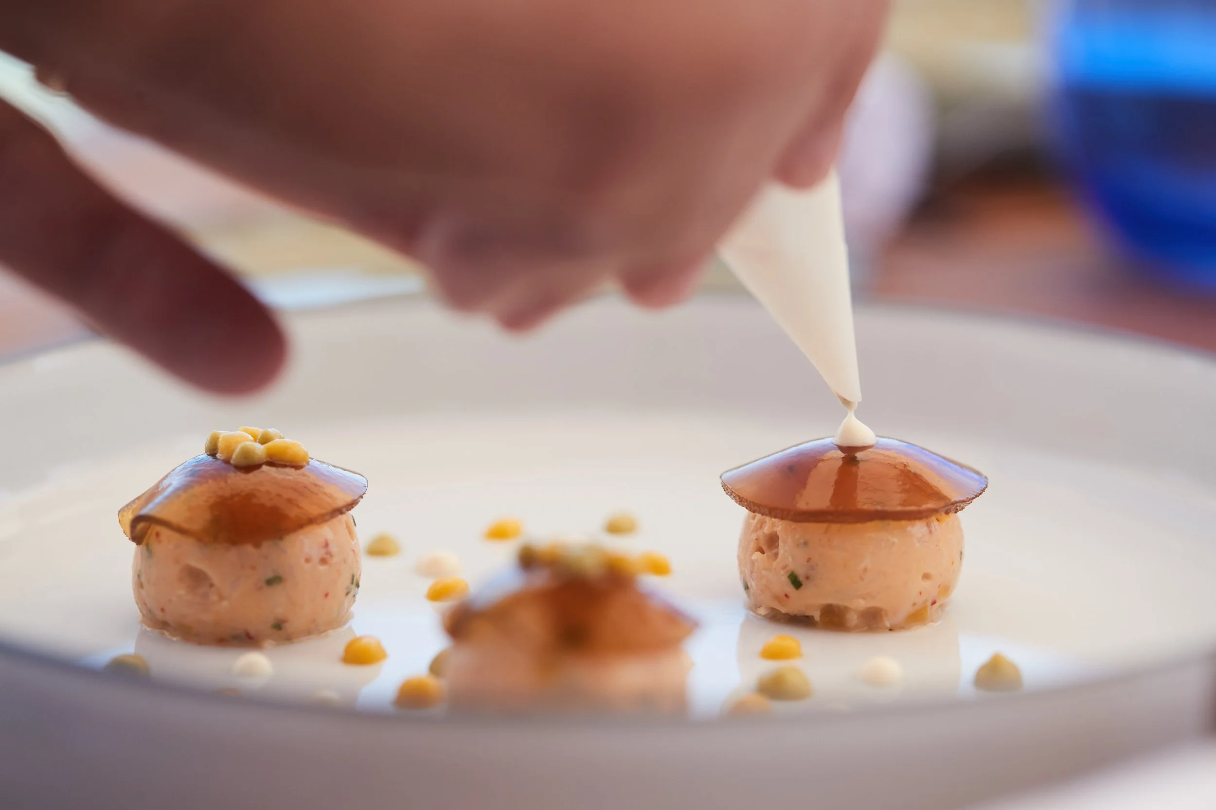 Chef decorates appetizer with piping bag, three small round food portions with brown sauce and small yellow and white garnishes on white plate.