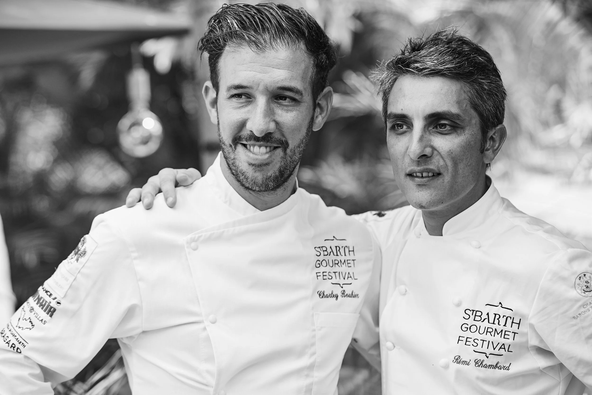 Two male chefs in white uniforms at the Sbarth Gourmet Festival, with one arm around each other's shoulders, standing outdoors.