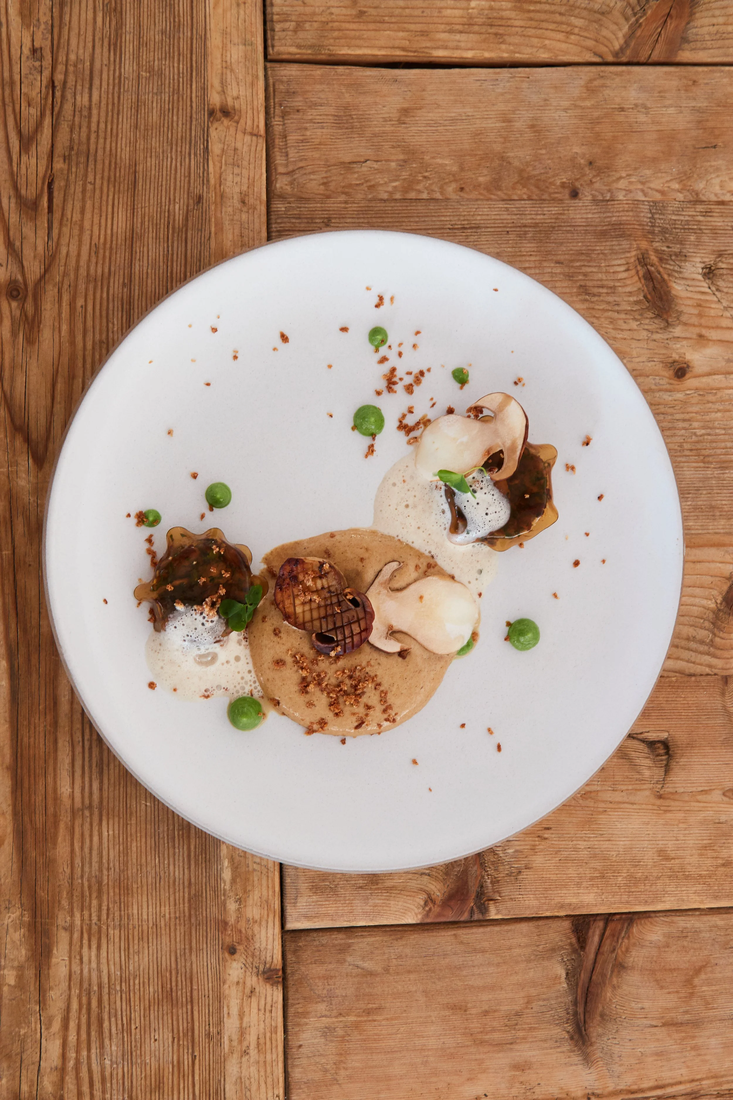 Gourmet plated dessert with foam, chocolate accents, green spheres, and edible flowers on a white plate, placed on a wooden surface.