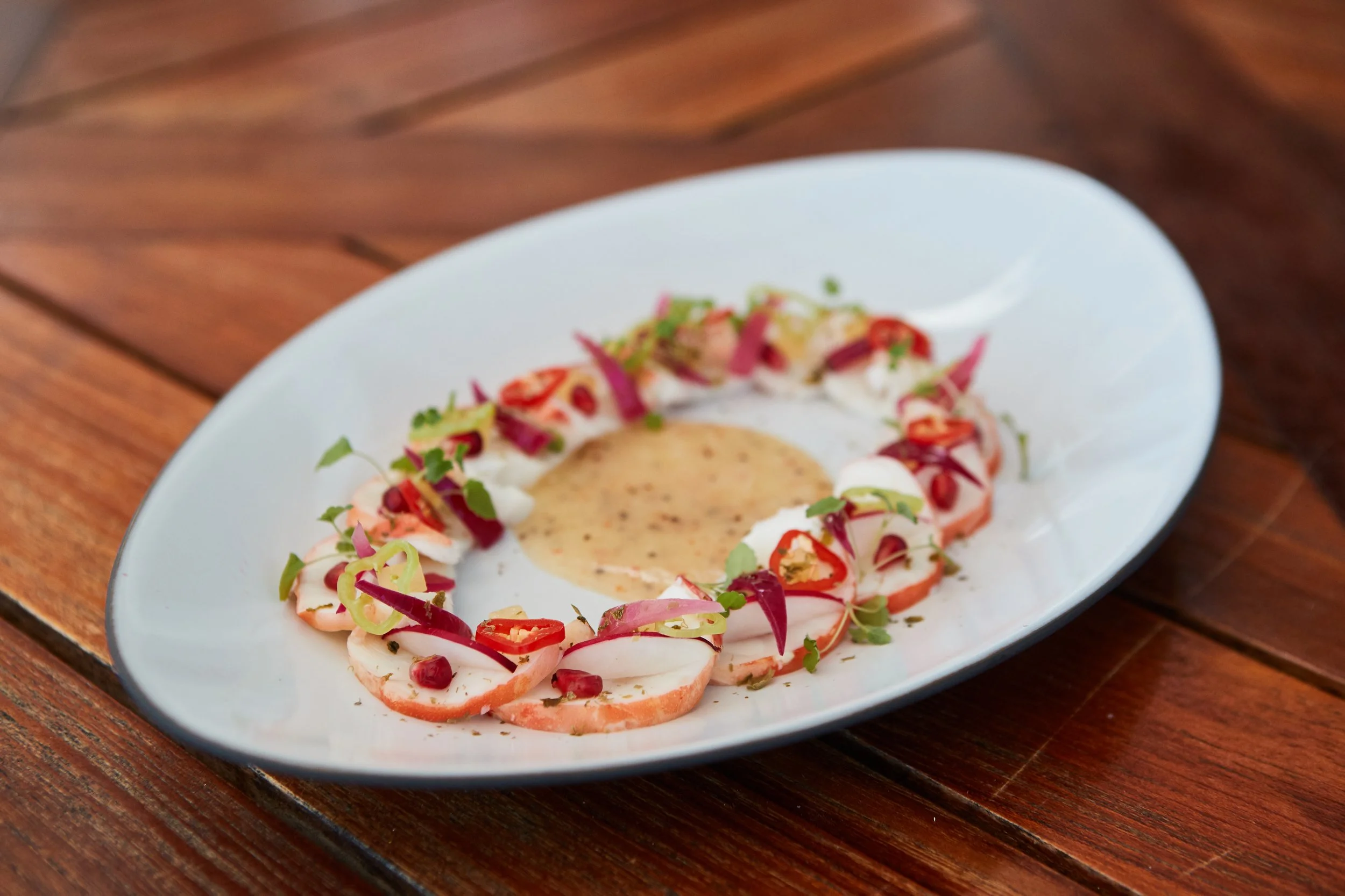 A plate of sliced tomatoes topped with herbs, arranged in a circle around a central scoop of creamy sauce with herbs, on a wooden table.