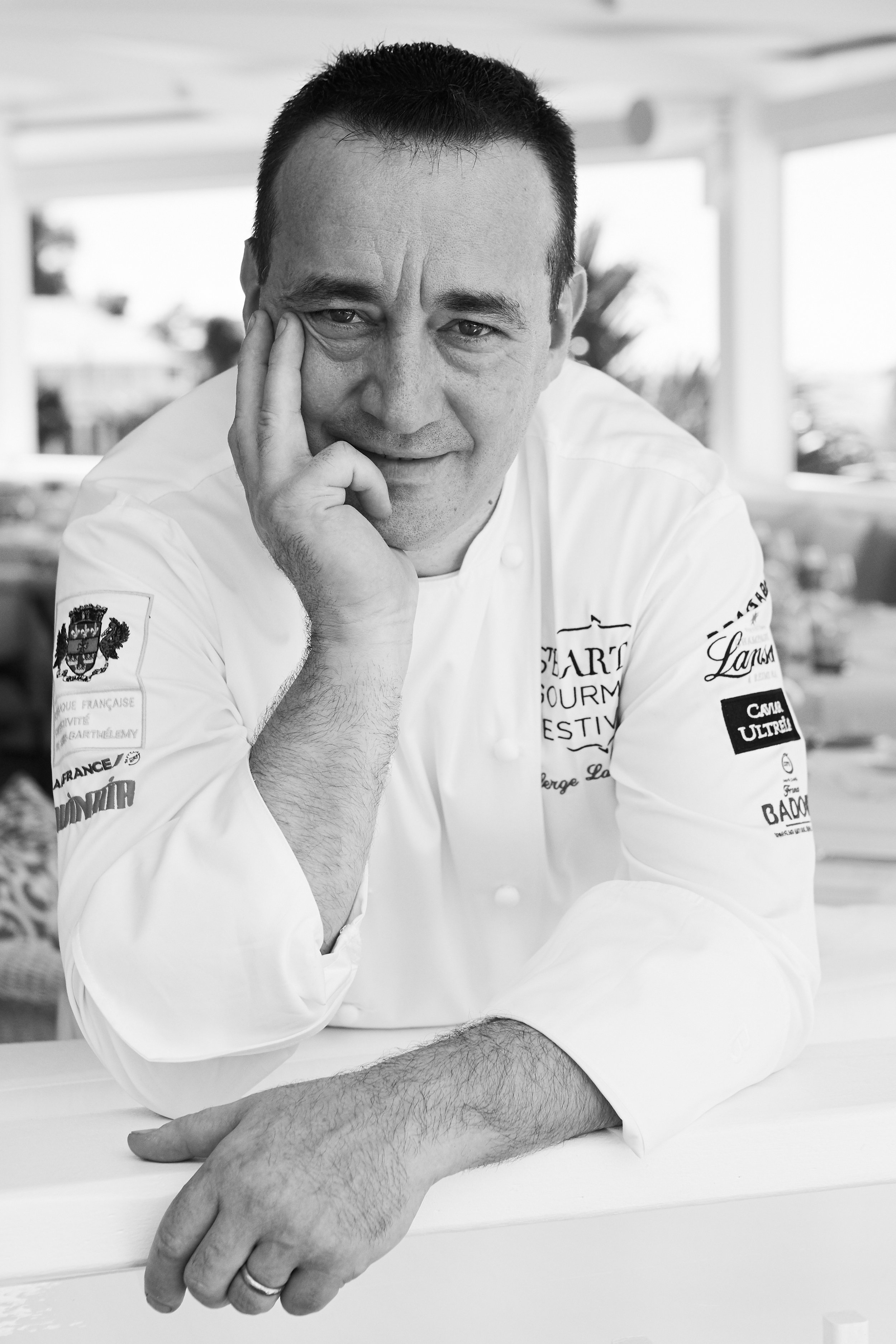 Black and white photo of a male chef resting his chin on his left hand, smiling slightly, in an indoor setting with bright background. He is wearing a white chef coat with logos and patches.