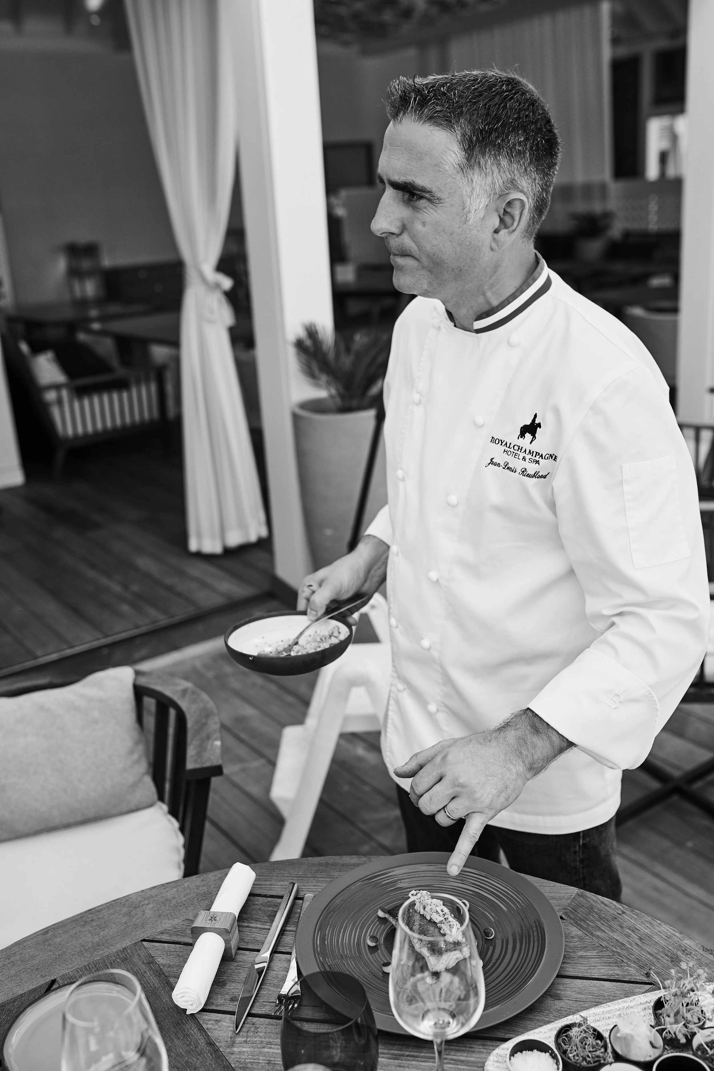 A chef in a white uniform with a logo and text, standing at a dining table with a plate and a glass, holding a small bowl with food, in a restaurant setting.