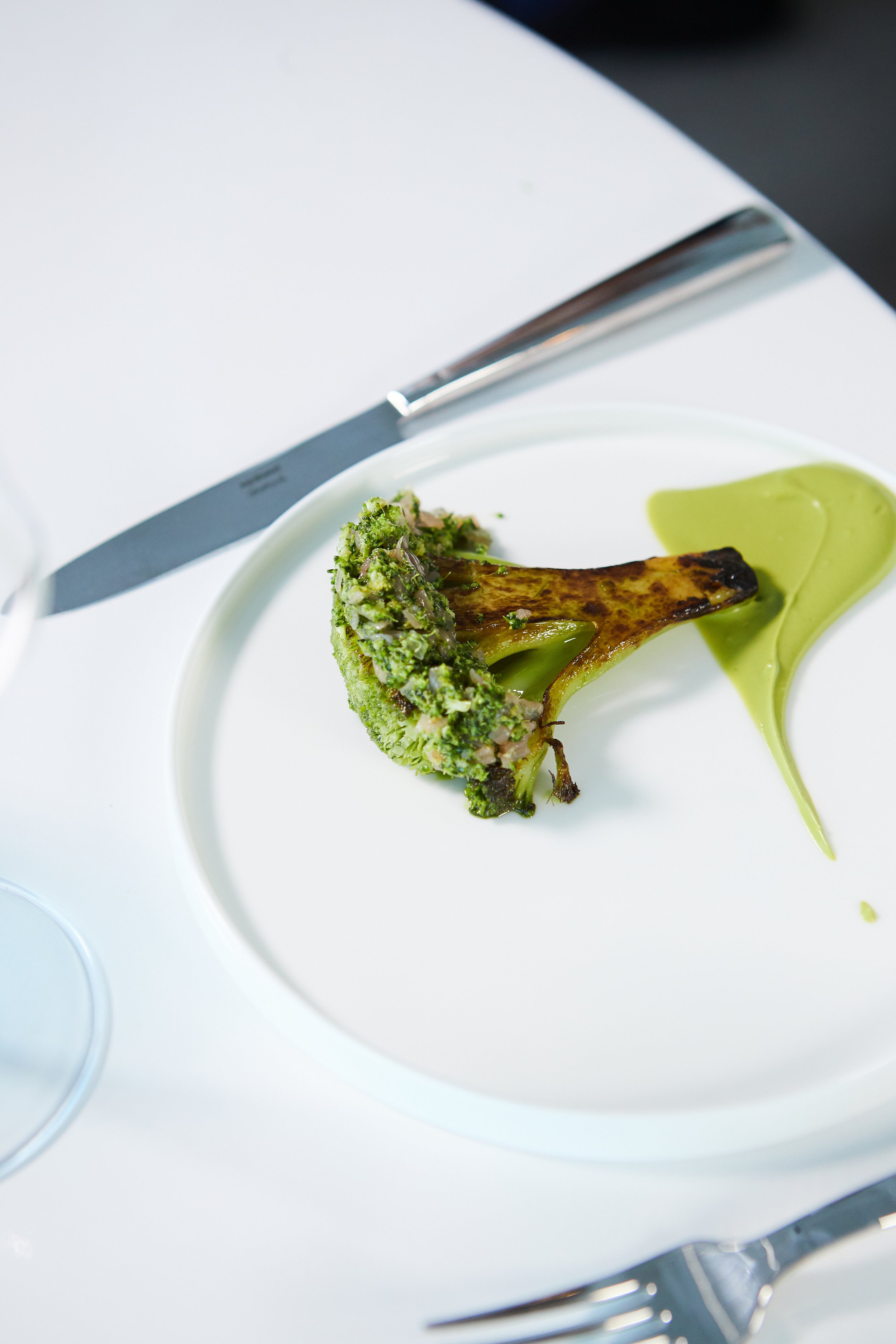 A white plate with a roasted broccoli floret and a smear of green sauce, placed on a white table with a knife and fork nearby.
