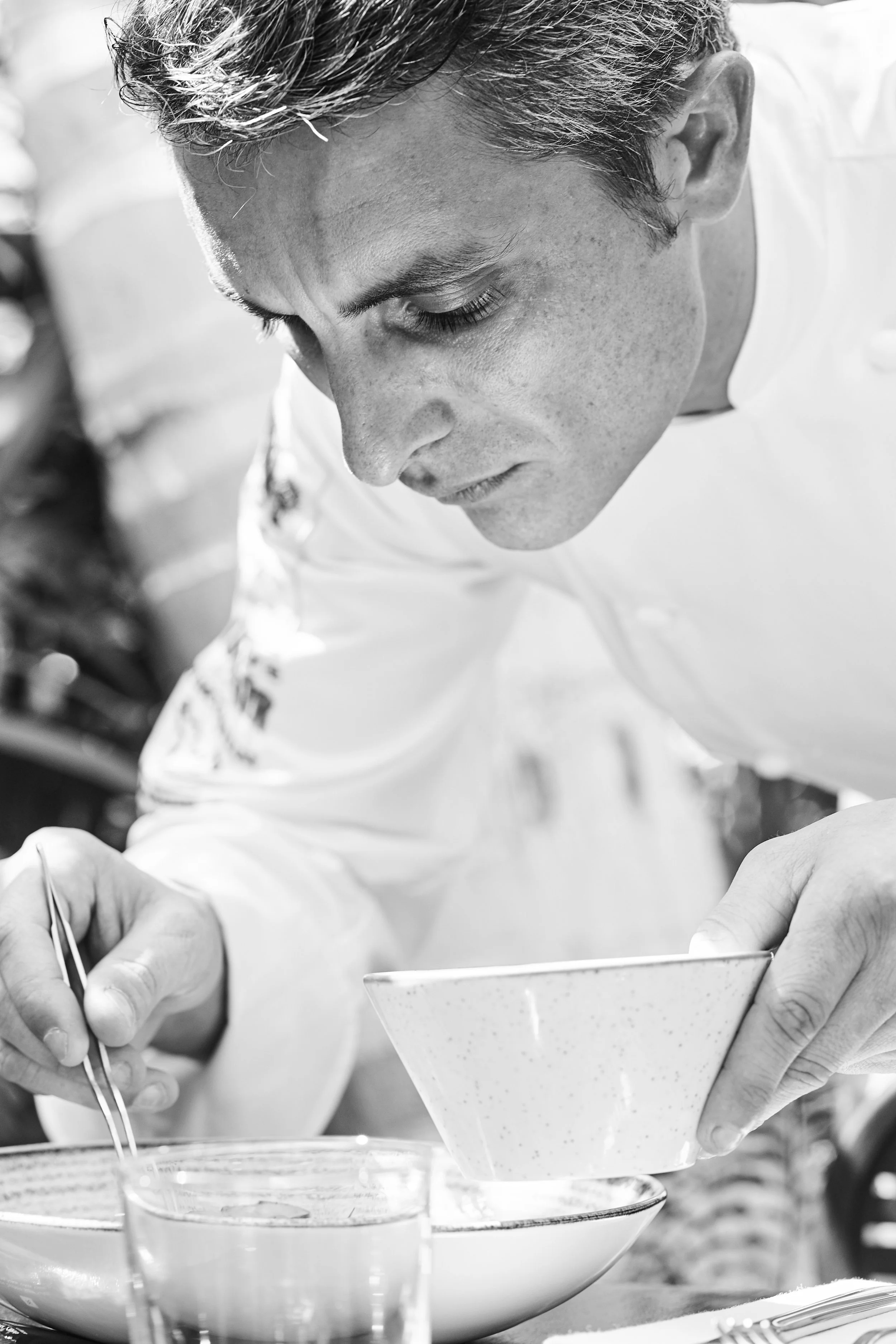 A man in a white chef's jacket is carefully pouring sauce into a bowl at a table.