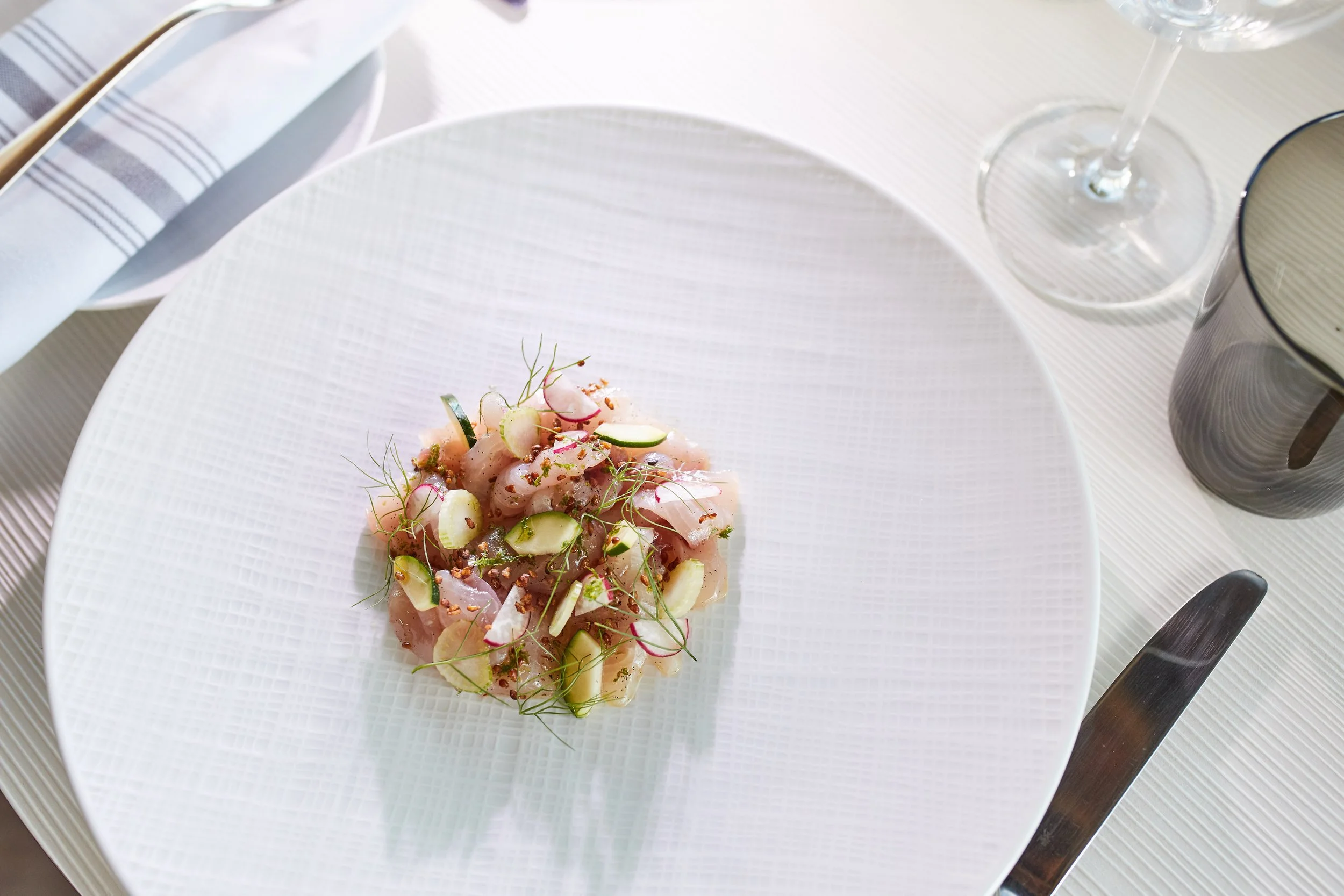 A plate of sashimi with thinly sliced fish garnished with microgreens, cucumber, and sauce on a white textured plate, with a wine glass, knife, and cup on a white tablecloth.