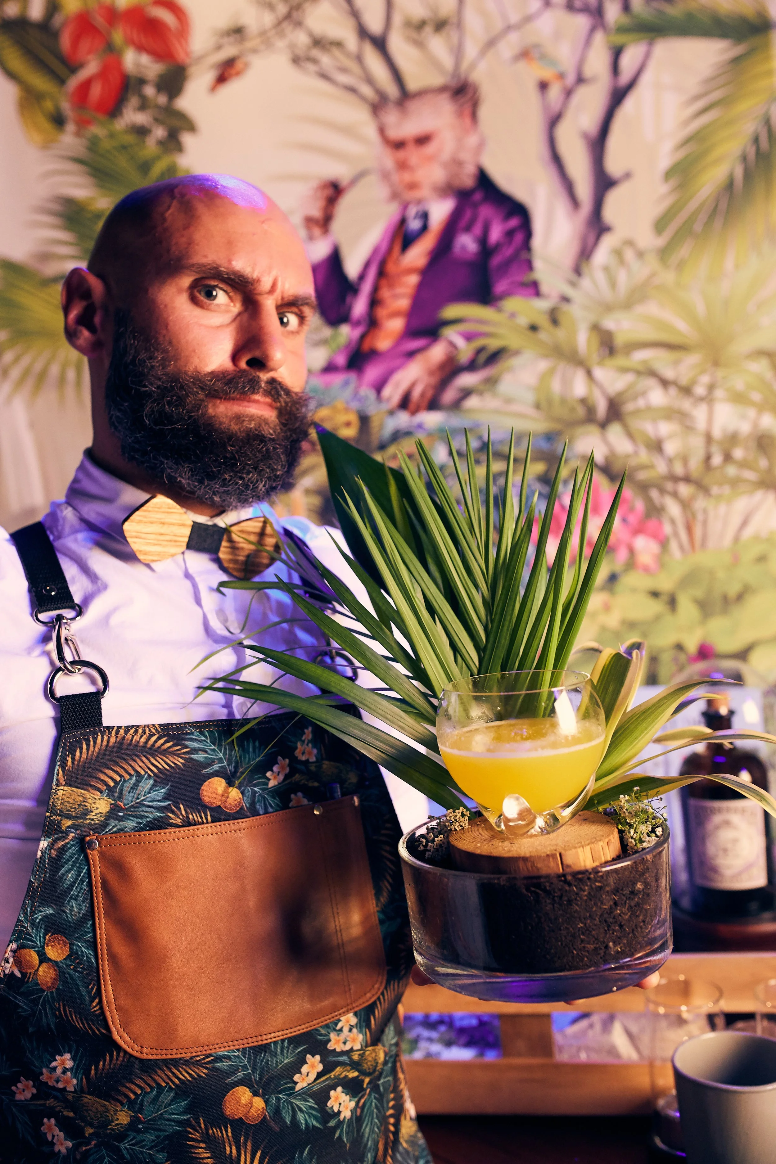 A bearded man wearing a white shirt, a floral apron with a brown pocket, and a wooden bowtie, holding a glass bowl with a yellow drink and tropical plants inside. Behind him is a colorful mural of a monkey in a purple suit and surrounded by large tro