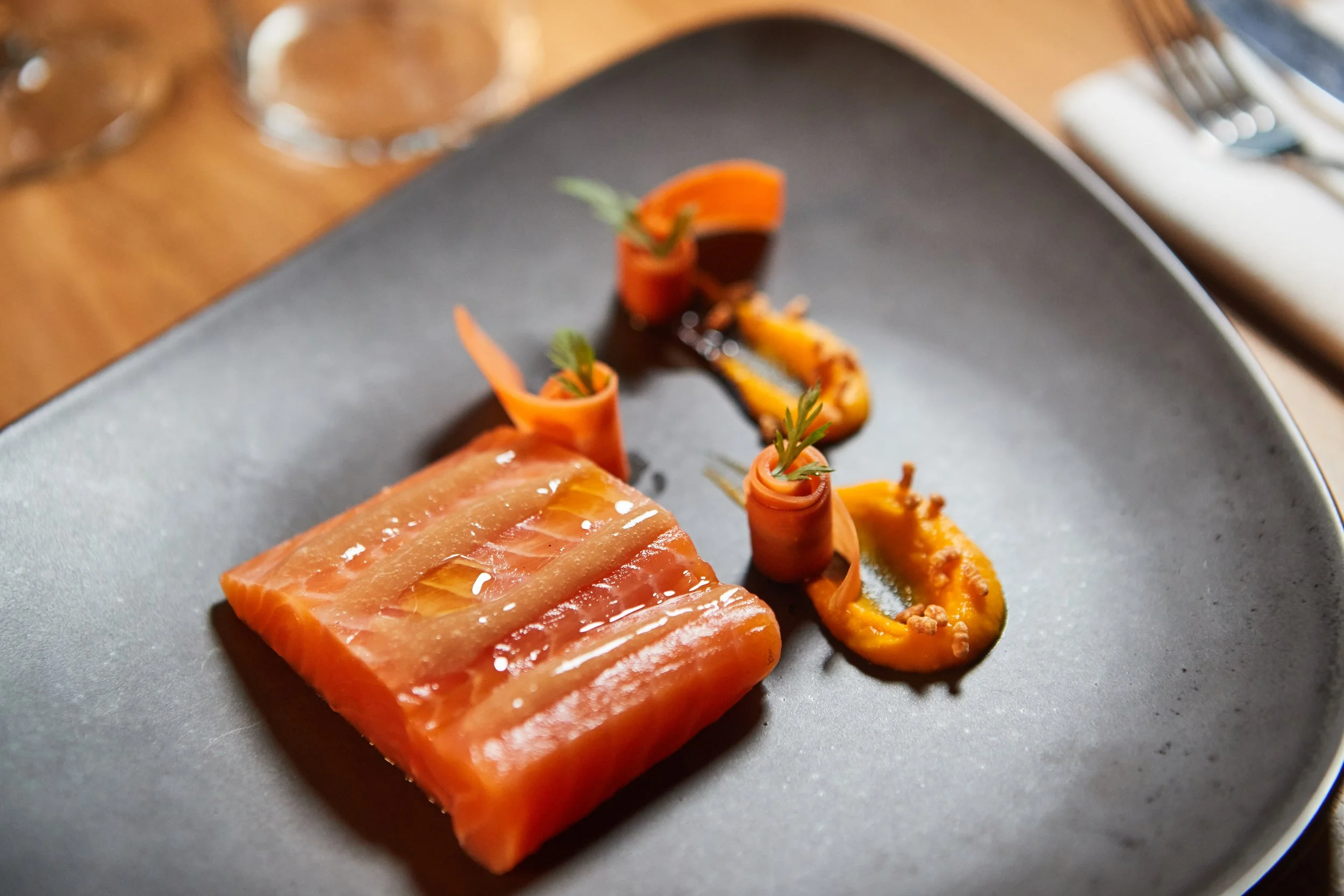 A plated dish featuring a large piece of smoked salmon with a glaze, accompanied by roasted squash and carrot curls garnished with herbs on a black plate.